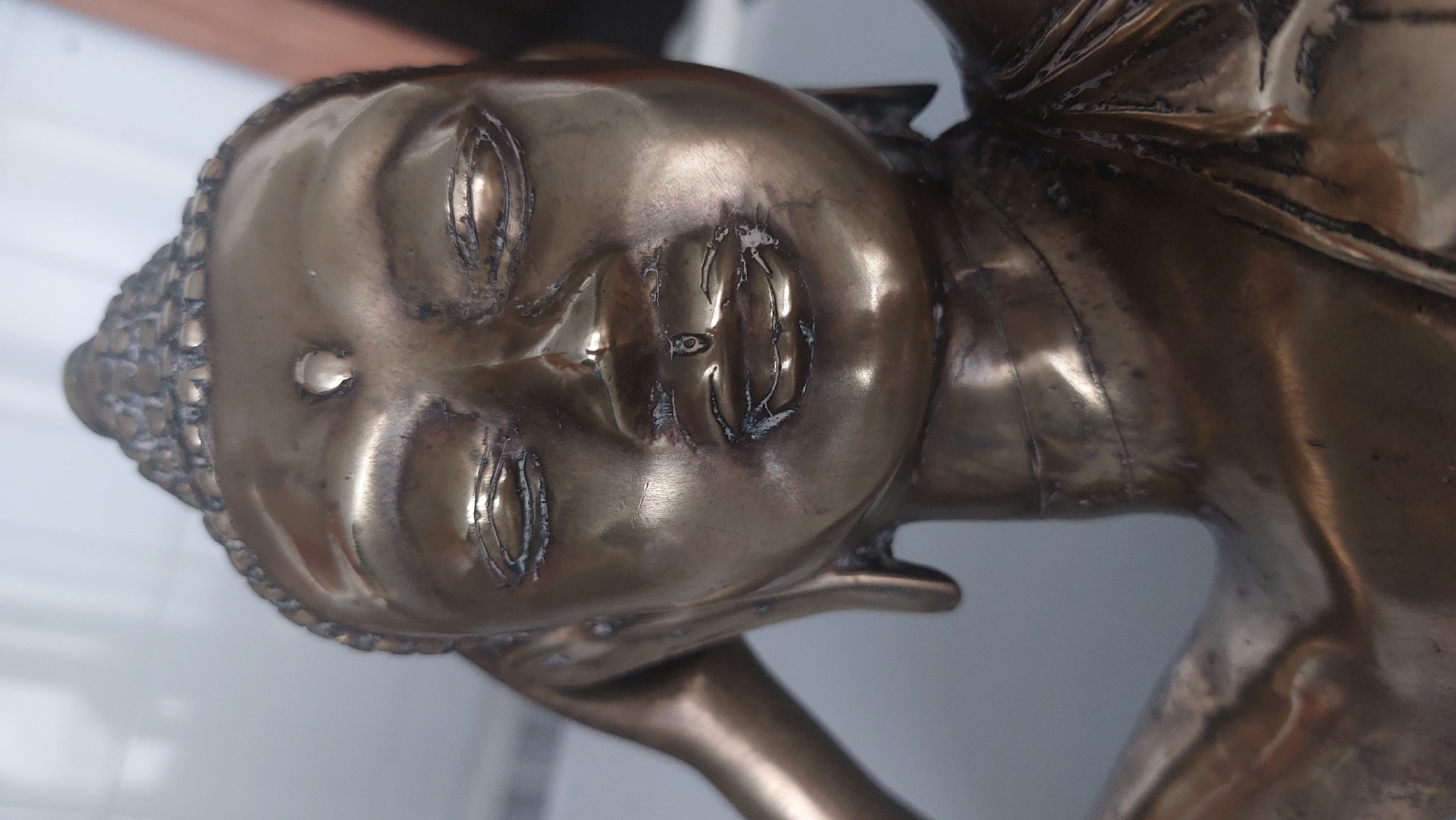 This is a close-up, vertical, eye-level shot of a bronze statue of the Buddha. The statue is detailed and appears aged, with some visible wear and imperfections. The Buddha's face is the primary focus, with eyes closed, a serene expression, and a distinctive urna (a decorative swirl or dot) on the forehead. The statue is illuminated from the front, creating reflections and highlights on the metallic surface. The background is blurred and out of focus, with indications of a light-colored wall or surface and possibly some darker elements to the right, suggesting an indoor setting. There is no discernible geographical information within the image to indicate its location in Andratx, Spain.
