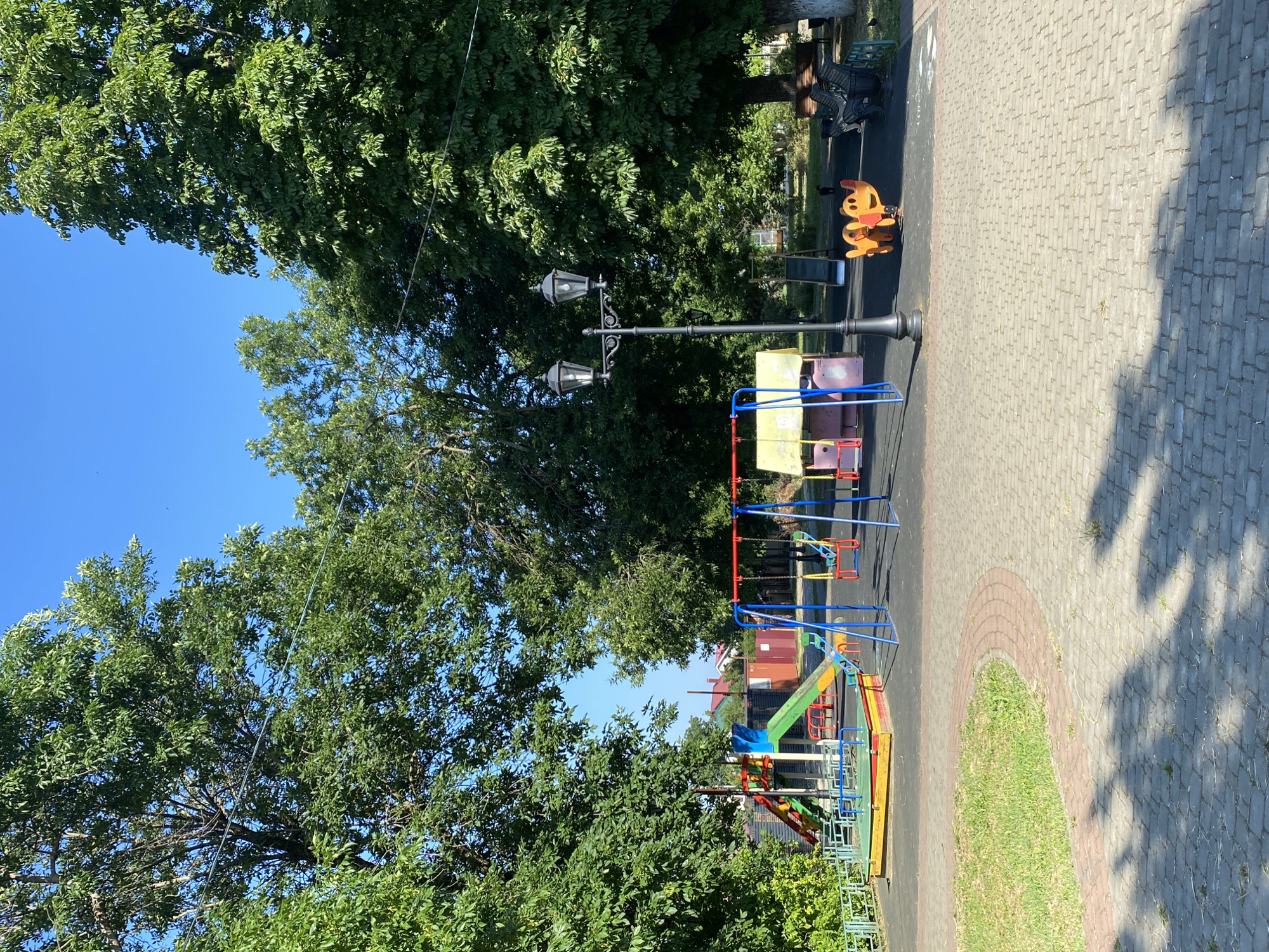 A sunny day at a public playground in Zapadny, Russia. The playground features a slide with a blue and green chute, a sandpit with yellow and red borders, and swings with red seats. There's also a yellow and pink playhouse and a bright orange spring rider shaped like a dog. A vintage-style double lamppost stands on a paved path, which curves around a patch of grass. In the background, mature trees with lush green foliage provide shade, and the clear blue sky is visible above. Several benches are positioned near the edge of the playground. Two individuals are seated on one of the benches in the far right of the image, observing the playground. The overall scene depicts a well-maintained and inviting recreational area.