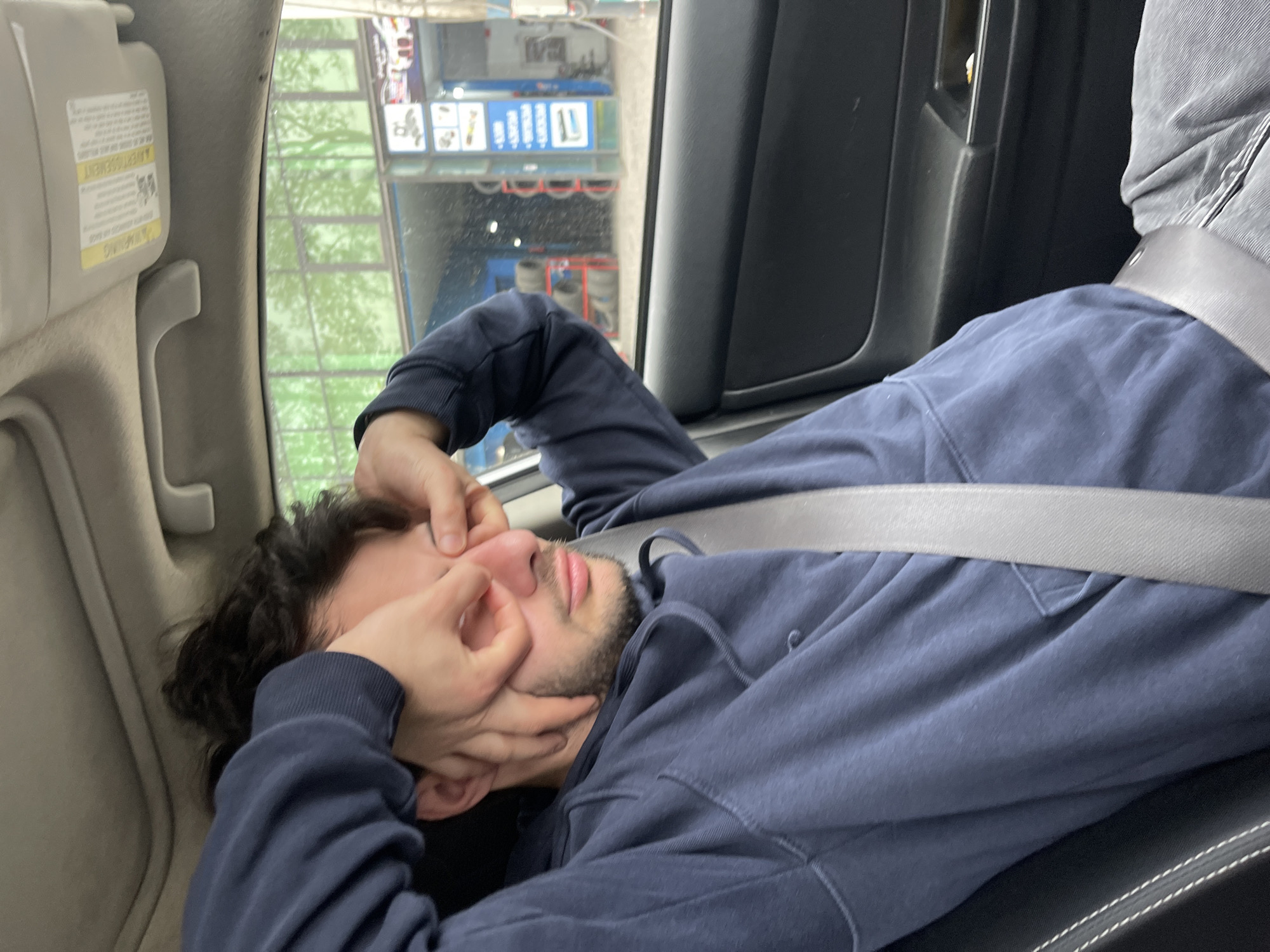 A man with dark curly hair and a beard is lying down in the backseat of a car wearing a blue hooded sweatshirt. He is wearing a seatbelt. He is pinching the bridge of his nose and appears to be in distress. Through the car window, the location appears to be Tbilisi, Georgia, based on the signage. The exterior appears to be a carwash.