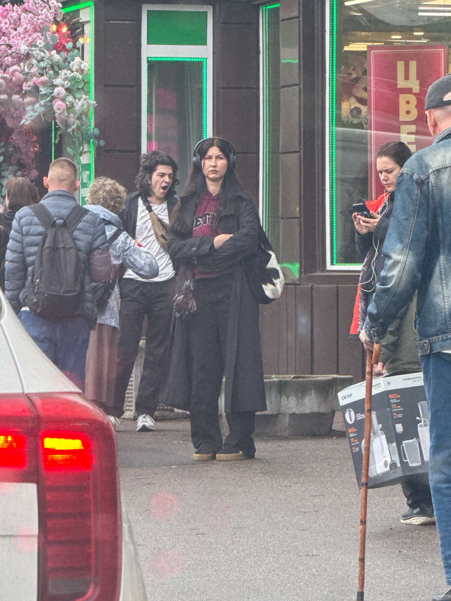 A group of people are standing on a sidewalk in front of a building with a dark facade and large windows. A young woman with long dark hair and headphones around her neck is in the foreground, looking forward with her arms crossed. To her left, a young man with curly dark hair is yawning. To the right of the woman, another woman is looking at her phone. Behind them, a man in a denim jacket is partially visible. In the foreground, the taillight of a car is visible, suggesting the scene is located on a street. The building appears to be a commercial establishment, with a pink sign in one of the windows displaying Cyrillic text. The overall atmosphere appears to be casual, with people waiting or passing by. The scene is likely in Saint Petersburg, Russia, given the architectural style and the presence of Cyrillic script.