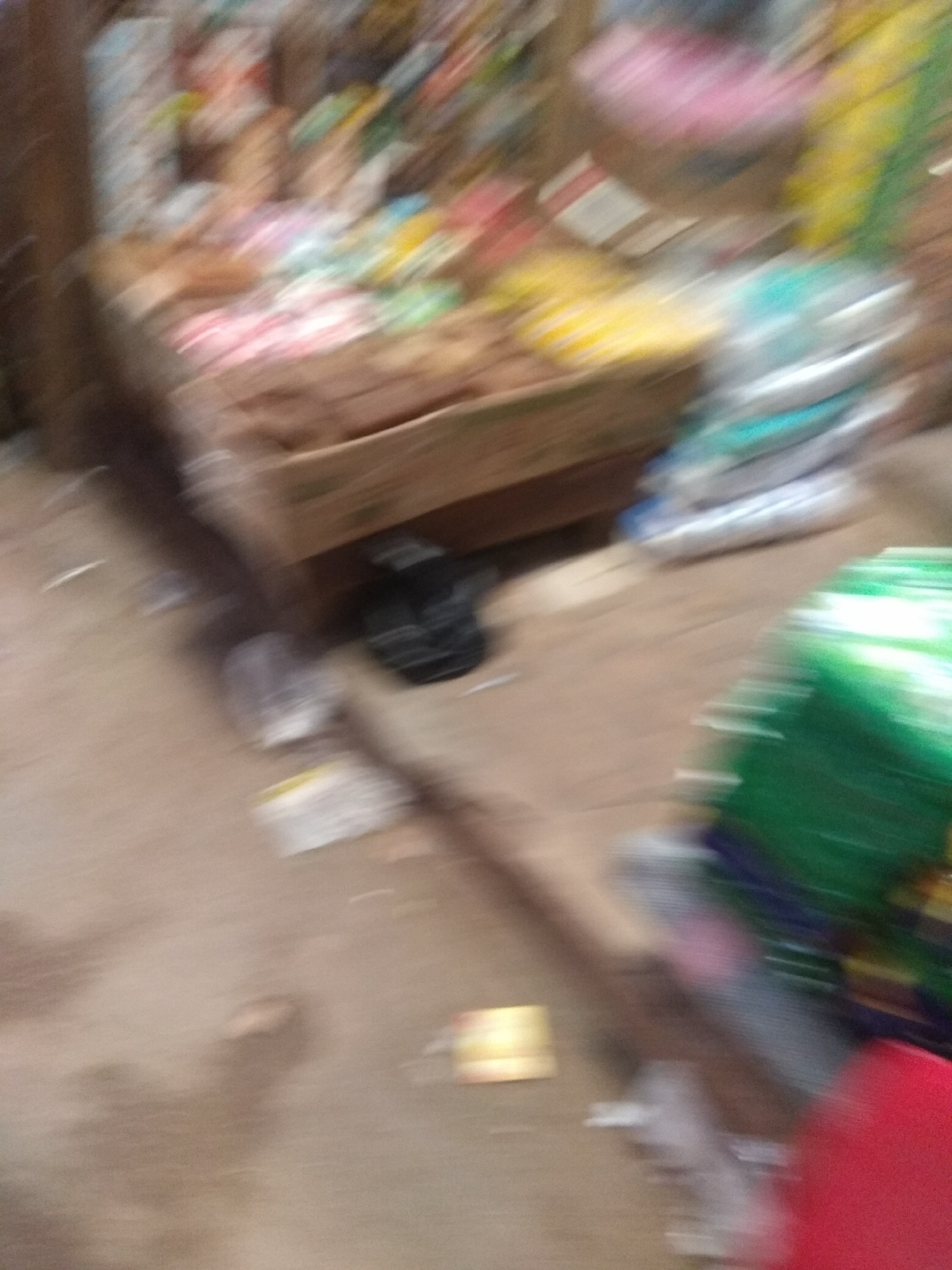 The image shows a blurred view of a market stall in Jalingo, Nigeria. The stall appears to be selling various goods, including packaged food items, snacks, and possibly household supplies. The items are stacked in wooden crates and on shelves. In the foreground, there is a pile of what looks like plastic bags, some green and some blue. To the left of the stall, there is a black object that resembles a shoe or a bag. The ground is dusty and appears to be made of packed dirt. The overall scene suggests a busy marketplace environment, typical of Nigerian cities. The image is very blurry due to motion, making it difficult to identify specific products or individuals.