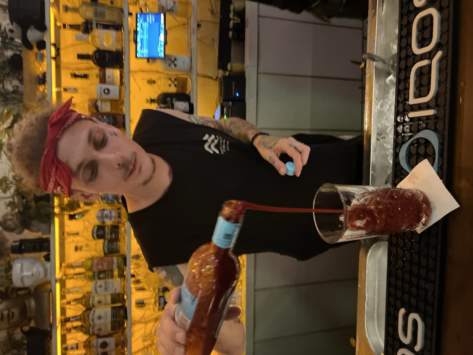A person wearing a black sleeveless t-shirt and a red bandana around their head is preparing a drink behind a bar. The person is holding a bottle filled with a red liquid and pouring it into a tall glass filled with ice and a red beverage. The person's left hand is holding a small blue object, possibly a cap or a garnish. Behind the person, shelves are stocked with various bottles of alcohol. In the background, a small blue screen displays some information. The bar counter has an ice well and is branded with the "IQOS" logo. The scene appears to be indoors, with warm yellow lighting illuminating the shelves. The location is not specified in the image or the provided context.