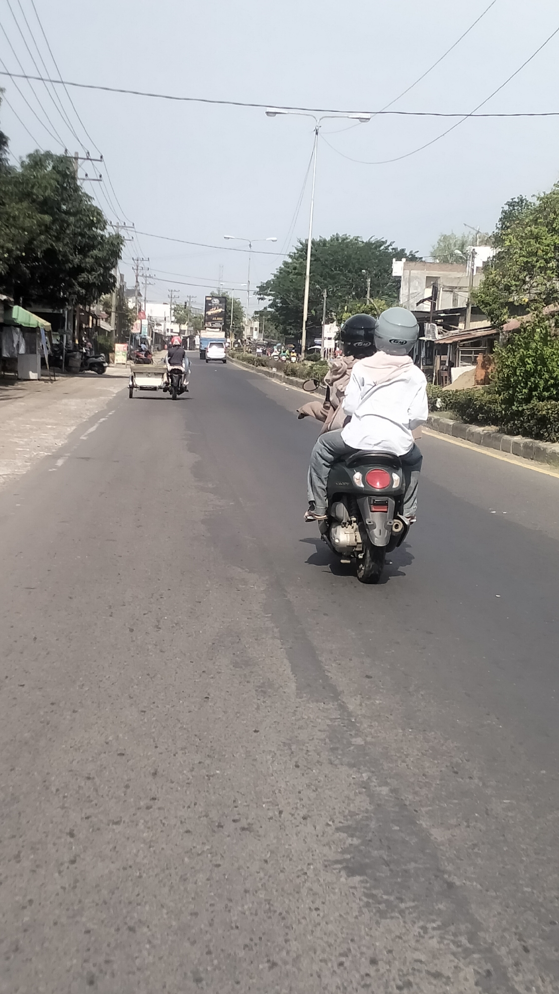 In Lhokseumawe, Indonesia, on a sunny day, a motorcyclist wearing a white shirt and jeans rides a dark-colored scooter, carrying a passenger behind them who is wearing a brown jacket and helmet. They are on a paved road, possibly a main street, with trees and buildings lining the sides. Other vehicles, including another motorcycle with a trailer, cars, and more motorcycles, are visible in the distance on the same road. The environment appears to be a moderately developed urban or suburban area. The sky is clear and bright.