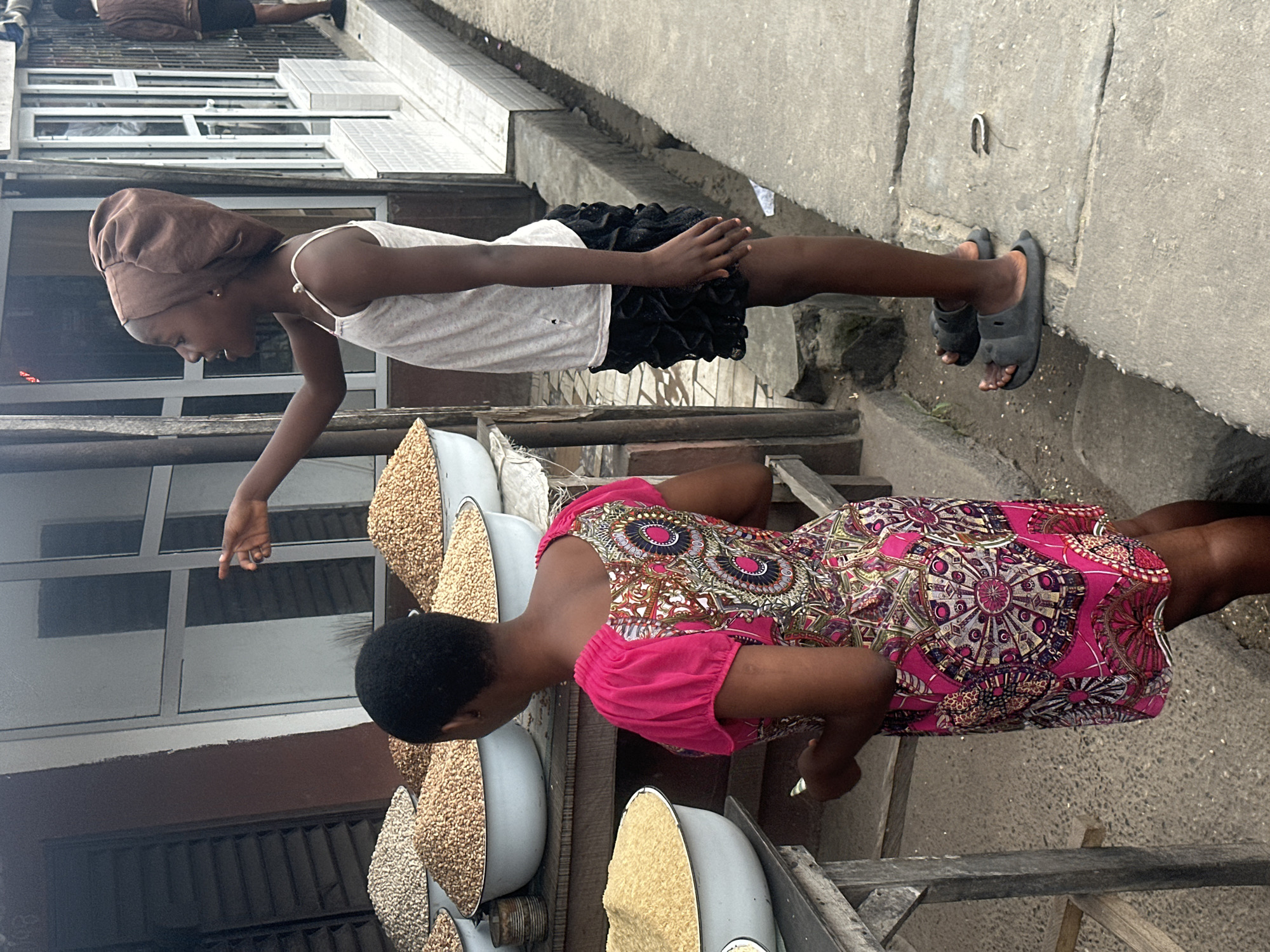 In Badagry, Nigeria, two young girls are positioned in front of a market stall laden with sacks of grains. One girl, wearing a bright pink dress with a complex geometric pattern, stands with her back to the camera, facing a large bowl of yellow grain. She is holding a small object in her hand. The other girl, older and wearing a white tank top and a black ruffled skirt, stands to her right and faces the camera. She wears a brown headscarf and points with her left hand towards the grains. Her right hand is resting on her hip. The background shows a building with large windows and shuttered sections. The ground is paved with concrete slabs.