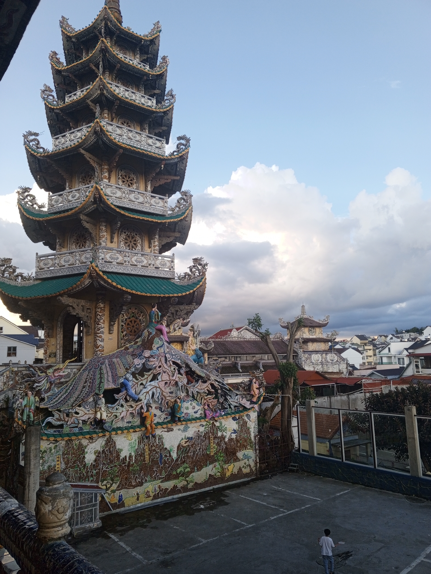 In Xuân Thành, Vietnam, an ornate, multi-tiered pagoda with intricate dragon carvings and green tiled roofs dominates the scene. The pagoda's exterior is adorned with colorful mosaics depicting figures and mythical creatures. In the foreground, a paved courtyard has a lone young person holding a white object, possibly a racket, facing away from the camera. The background reveals a dense cityscape with various buildings under a sky filled with dramatic clouds.
