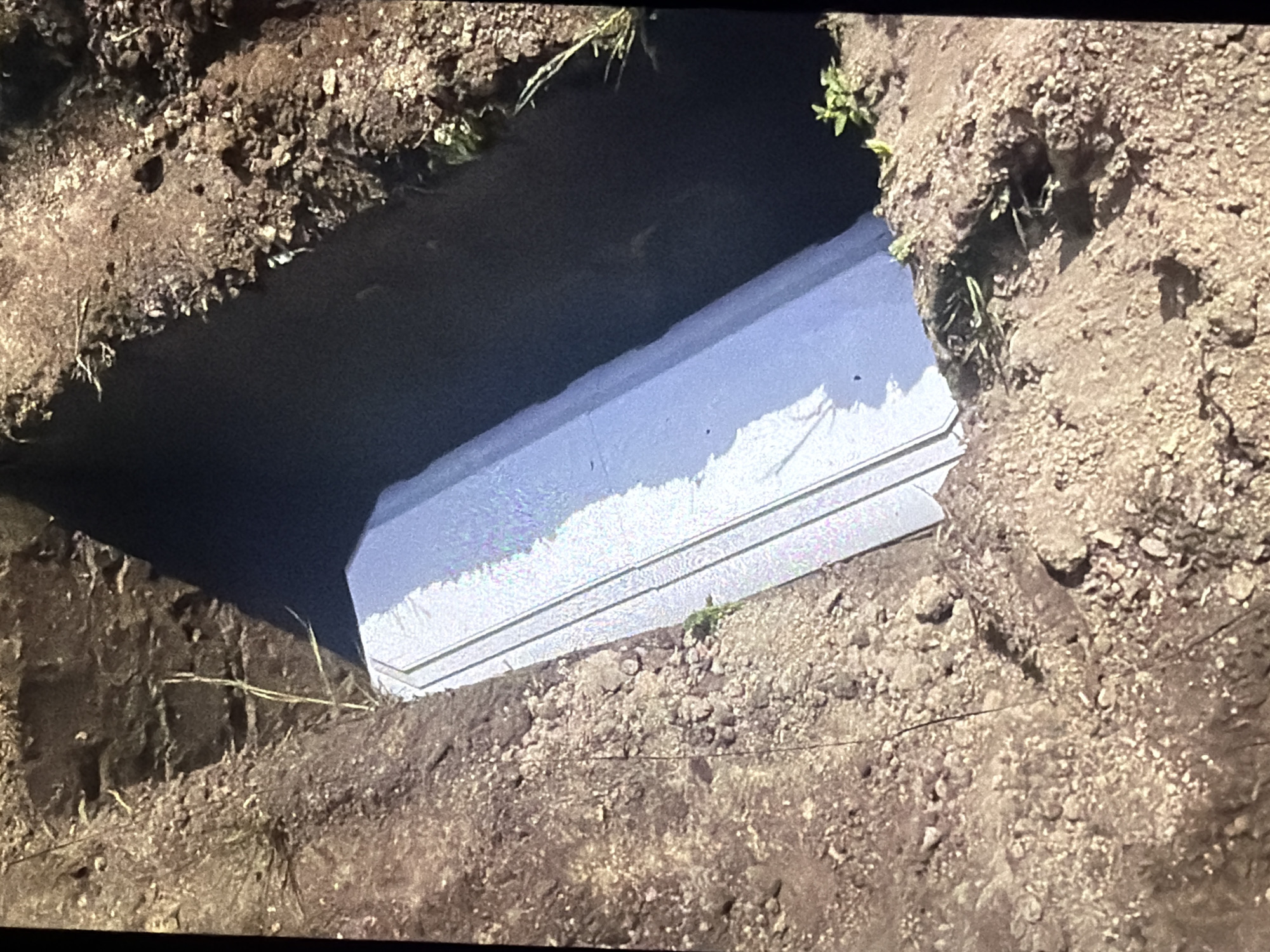 An aerial, close-up view shows a rectangular, light-colored coffin positioned inside a freshly dug grave. The grave walls are composed of dark, disturbed soil with visible rocks and sparse vegetation. The coffin appears to be partially embedded in the soil. The scene suggests a burial in an outdoor setting.