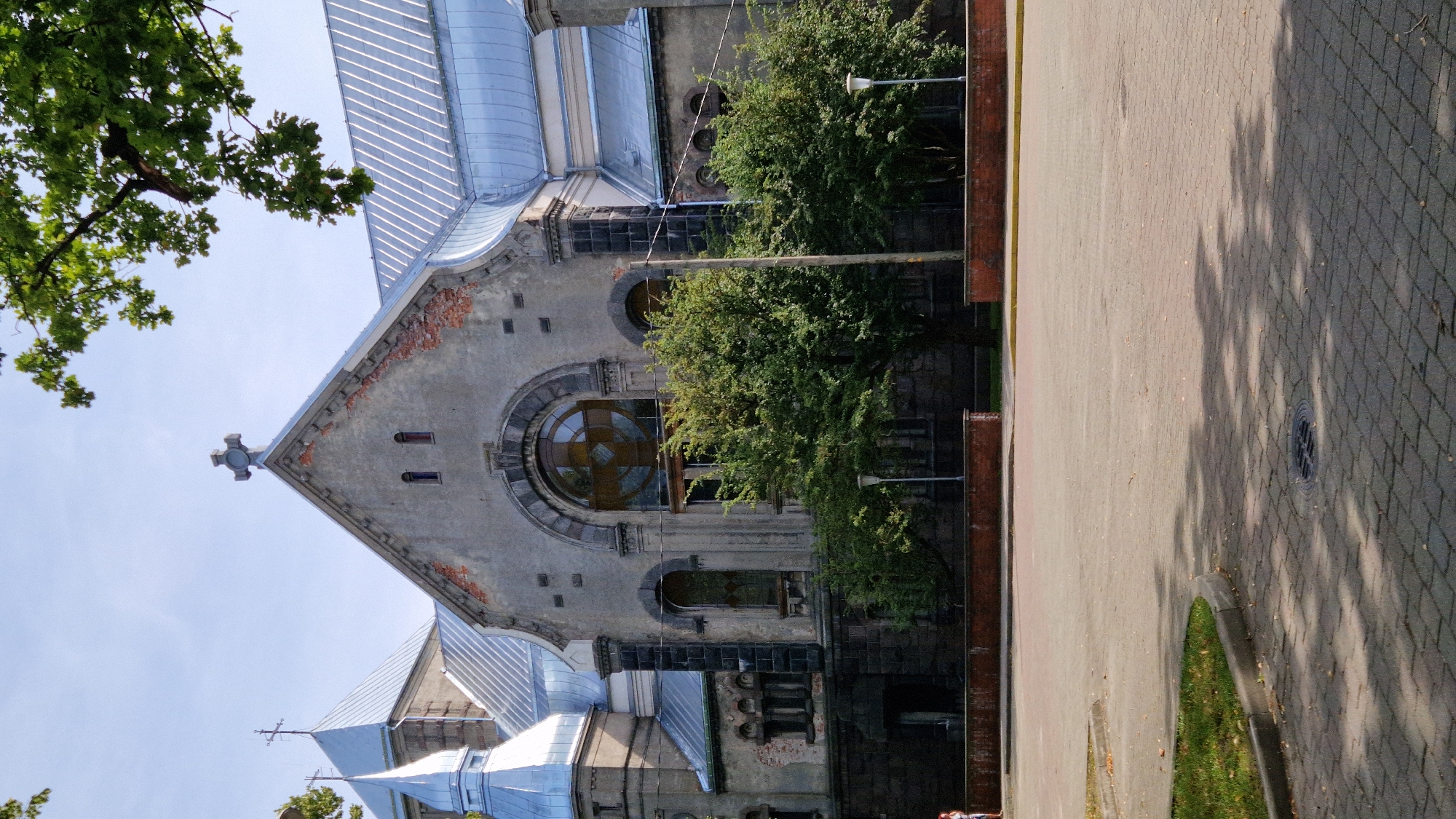 The image shows the exterior of St. Nicholas Orthodox Church in Liepaja, Latvia. The church is a large, stone building with a prominent gable roof and a smaller, conical roof to the left. The facade features a large, arched window with intricate geometric patterns and several smaller arched windows. A cross is visible on top of the main gable. The building is surrounded by trees and a paved area in front. The sky is clear and blue. A person is walking on the left side of the paved area.