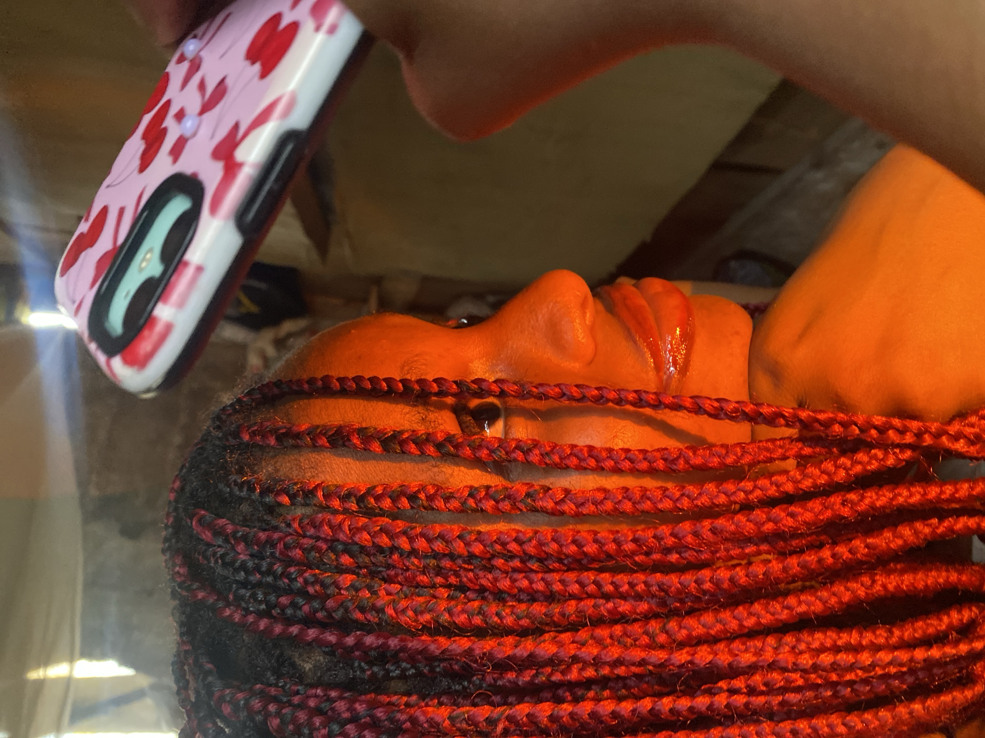 The image captures a close-up, upside-down view of a person's face and braided hair, illuminated by warm, orange light. The person's skin appears smooth and is covered in a sheen, suggesting sweat or oil. Their lips are slightly parted. The hair is styled in numerous, thick braids, a mix of black and vibrant red strands, which dominate the lower portion of the frame.
In the upper left corner, a portion of a phone case is visible. It is predominantly white with a pink pattern of what appears to be red hearts and small white dots. A sliver of the phone's screen is visible through the case, showing a teal-colored interface. The background is indistinct, appearing as a textured, possibly wooden surface.
The lighting suggests an indoor setting, potentially with artificial light sources casting the strong orange hue. The angle and focus of the image create an intimate and somewhat intense portrait, emphasizing the textures of the hair and skin. There are no discernible cues about the time of day or weather. The overall impression is of a moment captured during personal grooming or a selfie-taking session. There is no visible text in the image.