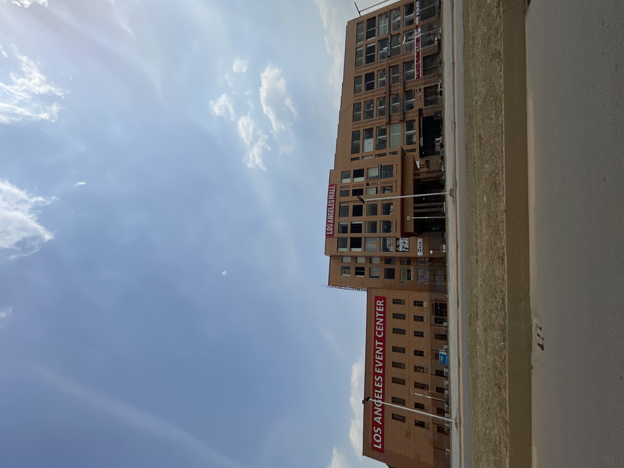 The image captures an exterior view of several multi-story buildings under a partially cloudy blue sky, depicting an urban setting in Abuja, Nigeria. The scene appears to be taken during the daytime, with ample natural light suggesting sunny weather.
The primary subjects are two light brown/tan colored buildings with numerous windows. The building on the left (as viewed when rotated correctly) features a large red sign vertically spelling "LOS ANGELES EVENT CENTER". The building further to the right has another prominent red vertical sign that reads "LOS ANGELES MALL". Other smaller signs are visible on the buildings, including "BAKAN GIZO PHARMACY & STORES", "EXCLUSIVE GALLERY", "Deye", and partially visible "azusa HOTEL".
In the foreground, a gray asphalt or concrete road runs parallel to the buildings. A strip of dry, light-colored ground or sparse vegetation separates the road from the building facades. The sky is largely clear blue with scattered white clouds, some appearing wispy. No people or active events are discernible in the photograph.