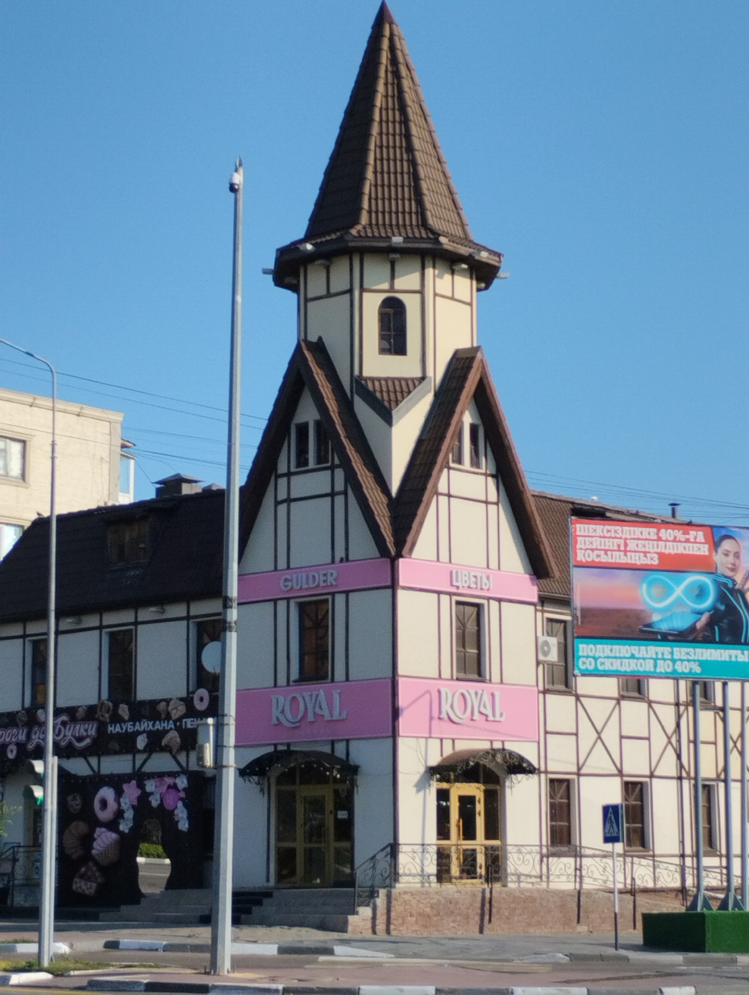 The image shows a two-story building with a distinctive Tudor-style architecture, featuring a tall, pointed roof and half-timbered detailing. The building is painted in cream and brown colors. On the lower part of the building, there are signs with Cyrillic text. One sign reads "ROYAL" in pink lettering. Another sign, visible on a billboard to the right of the building, displays an advertisement with a woman and a graphic of an infinity symbol, and text in Cyrillic stating "ПОДКЛЮЧАЙТЕ БЕЗЛИМИТЫ СО СКИДКОЙ ДО 40%". The building appears to be a commercial establishment, possibly a cafe or bakery, given the display of donuts on the left side. The setting is an urban environment, with a clear blue sky in the background and a street with road markings and a lamppost in the foreground. The image was taken in Kostanay, Kazakhstan. There are no people clearly visible in the image. The scene is brightly lit, suggesting it was taken during the daytime.