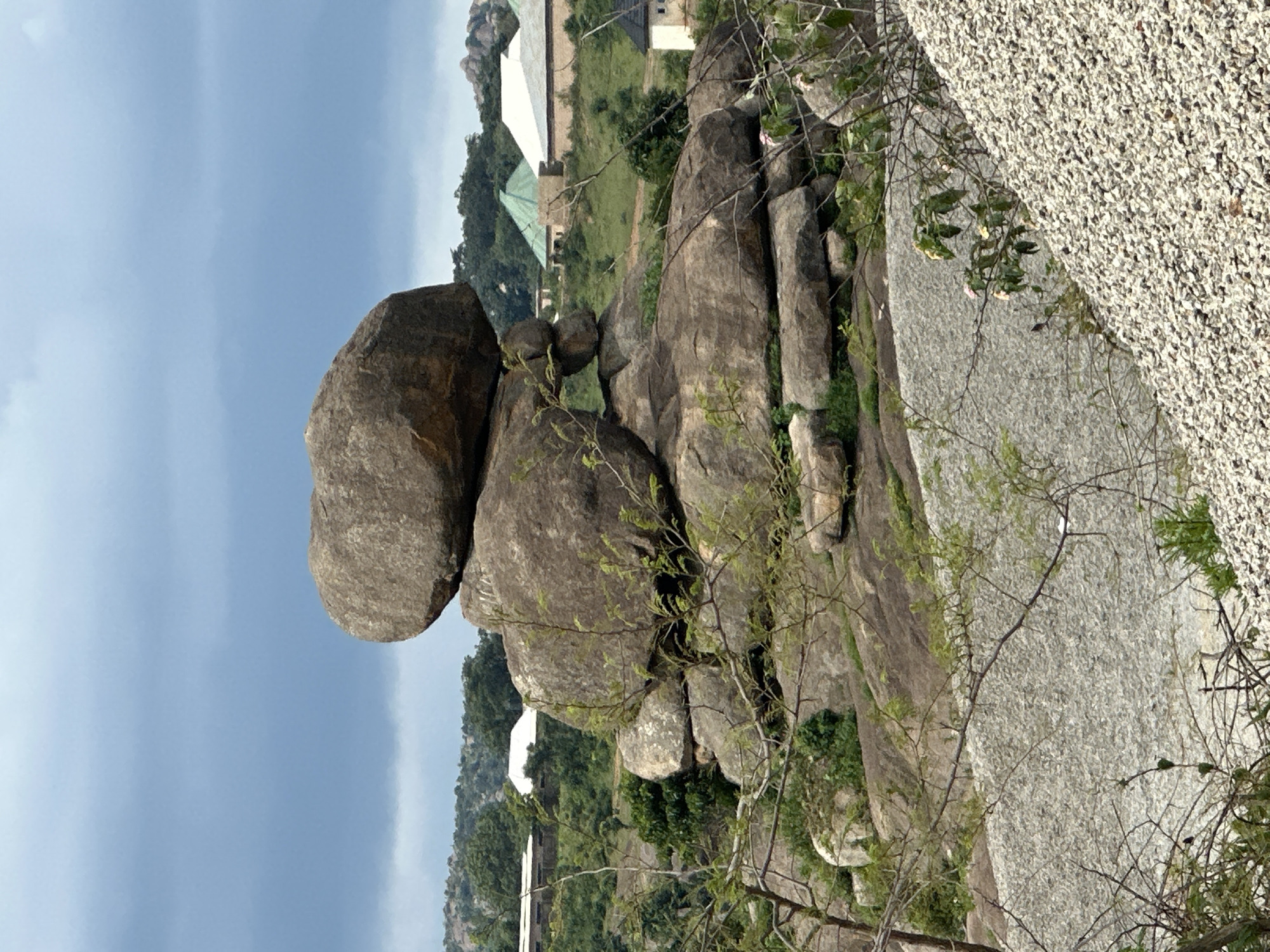 The image depicts a scenic landscape in Jos, Nigeria, dominated by a large, unusual rock formation. The rock appears to be composed of several massive, rounded boulders stacked precariously on top of each other. The top boulder is particularly prominent, resembling an upside-down mushroom. Vegetation, including trees and shrubs, grows around and on the rocks, suggesting a natural, possibly untamed environment. In the background, several buildings with distinct architectural styles, including some with green and white roofs, are visible, indicating human habitation and development within the vicinity. The sky is overcast with gray clouds, hinting at potentially changing weather conditions. The foreground features a slope covered with gravel or small stones, and some branches of trees with sparse foliage. The overall impression is one of a rugged natural beauty integrated with human structures.