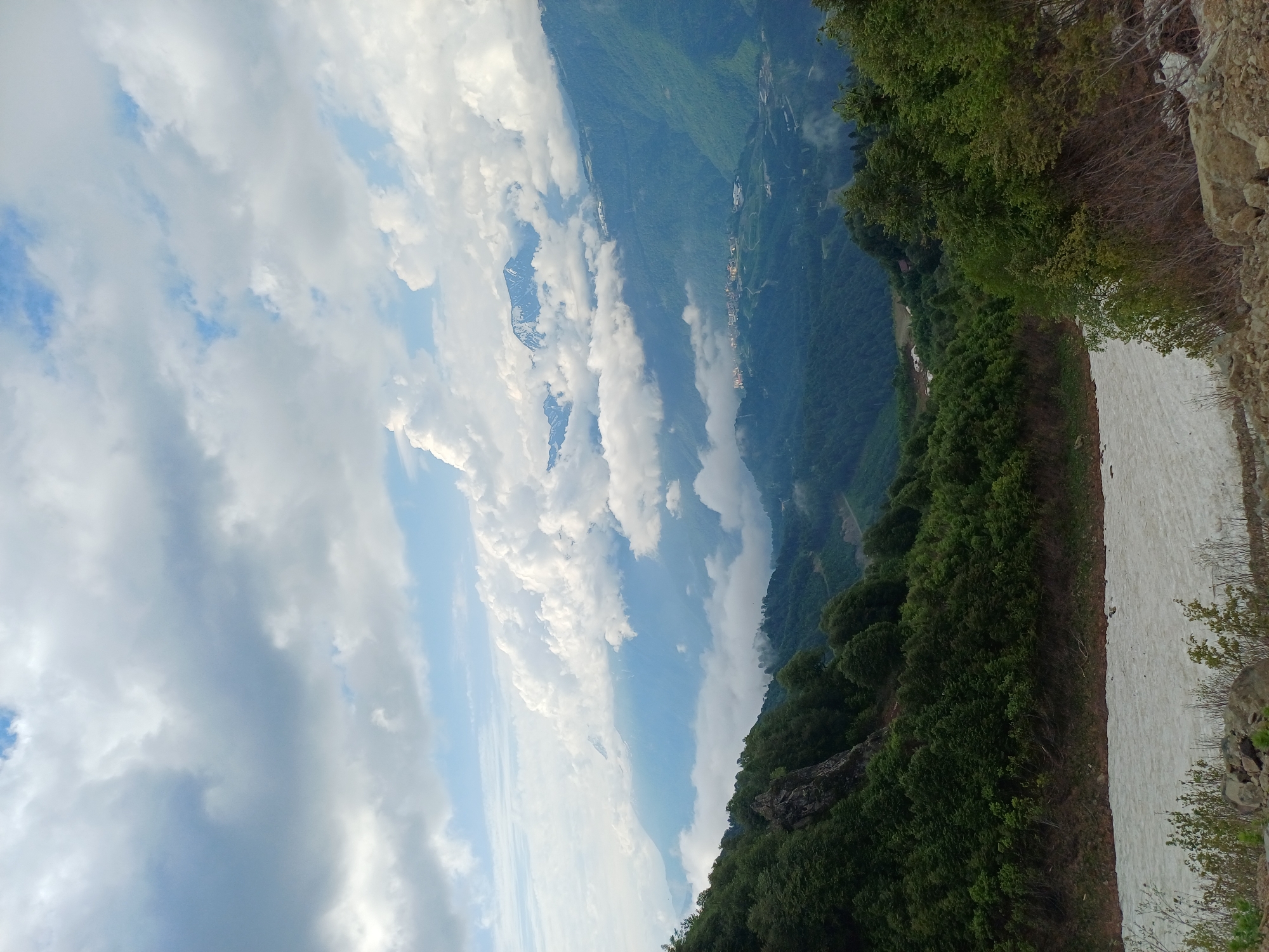 The image depicts a panoramic view of a mountainous landscape, likely in or around Estosadok, Russia. In the foreground, a patch of melting snow covers the ground, with some sparse vegetation and rocks. The midground is dominated by a steep, heavily wooded slope of green trees. Further in the background, layers of mountains recede into the distance, some shrouded in clouds. A small settlement with buildings is visible on a lower mountain slope, and the peaks of distant snow-capped mountains can be seen peeking through the clouds. The sky is filled with a dramatic display of white, puffy clouds against a light blue sky. The overall scene conveys a sense of natural beauty and the vastness of the mountain terrain.