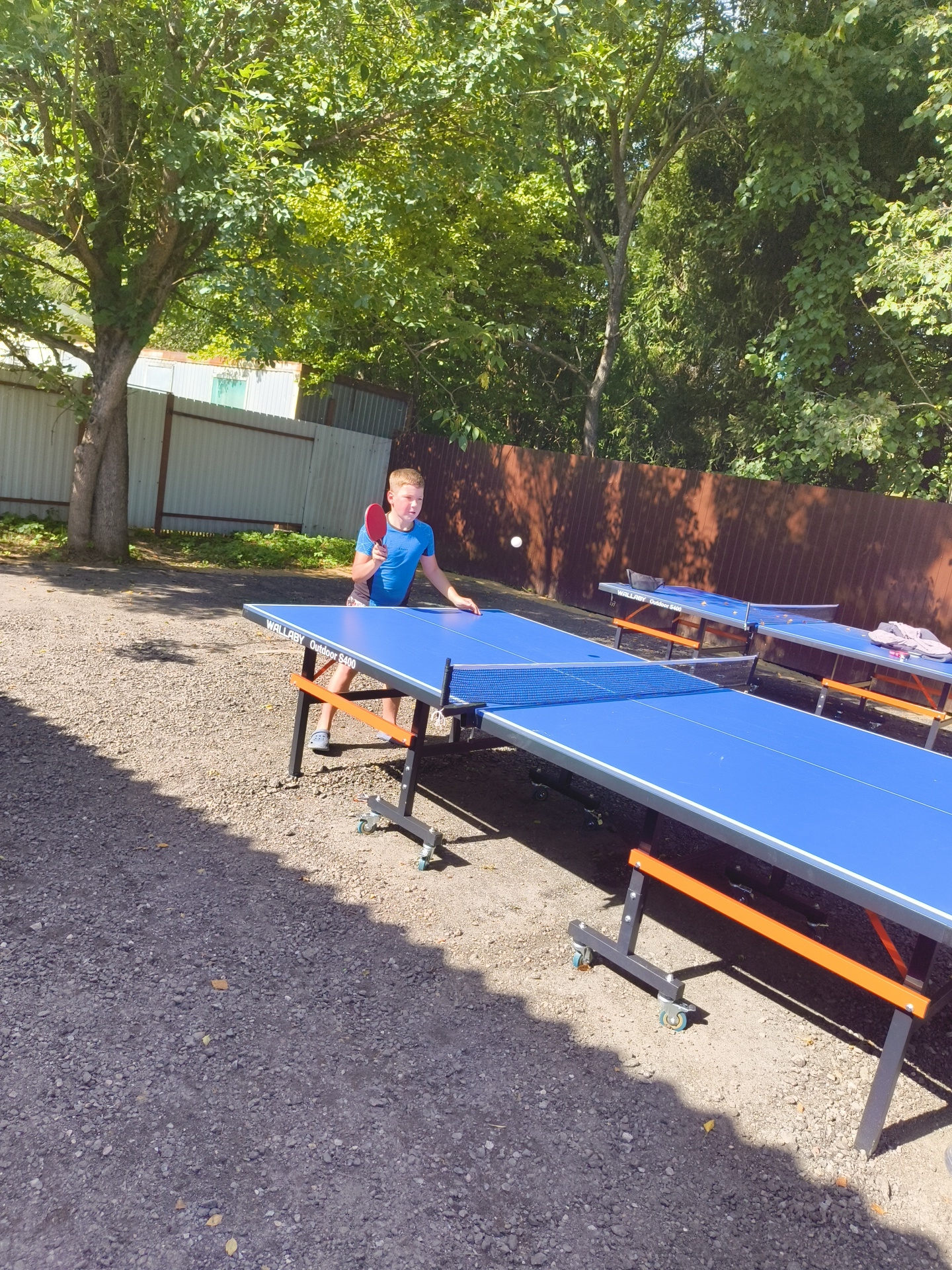 A boy is playing ping pong outside in a yard. He is standing next to a blue ping pong table and holding a red paddle. A ping pong ball is in the air. The yard is covered in gravel and surrounded by trees. There are fences in the background. The image appears to be taken in the city of Pravdist-2 SNT, Russia.
