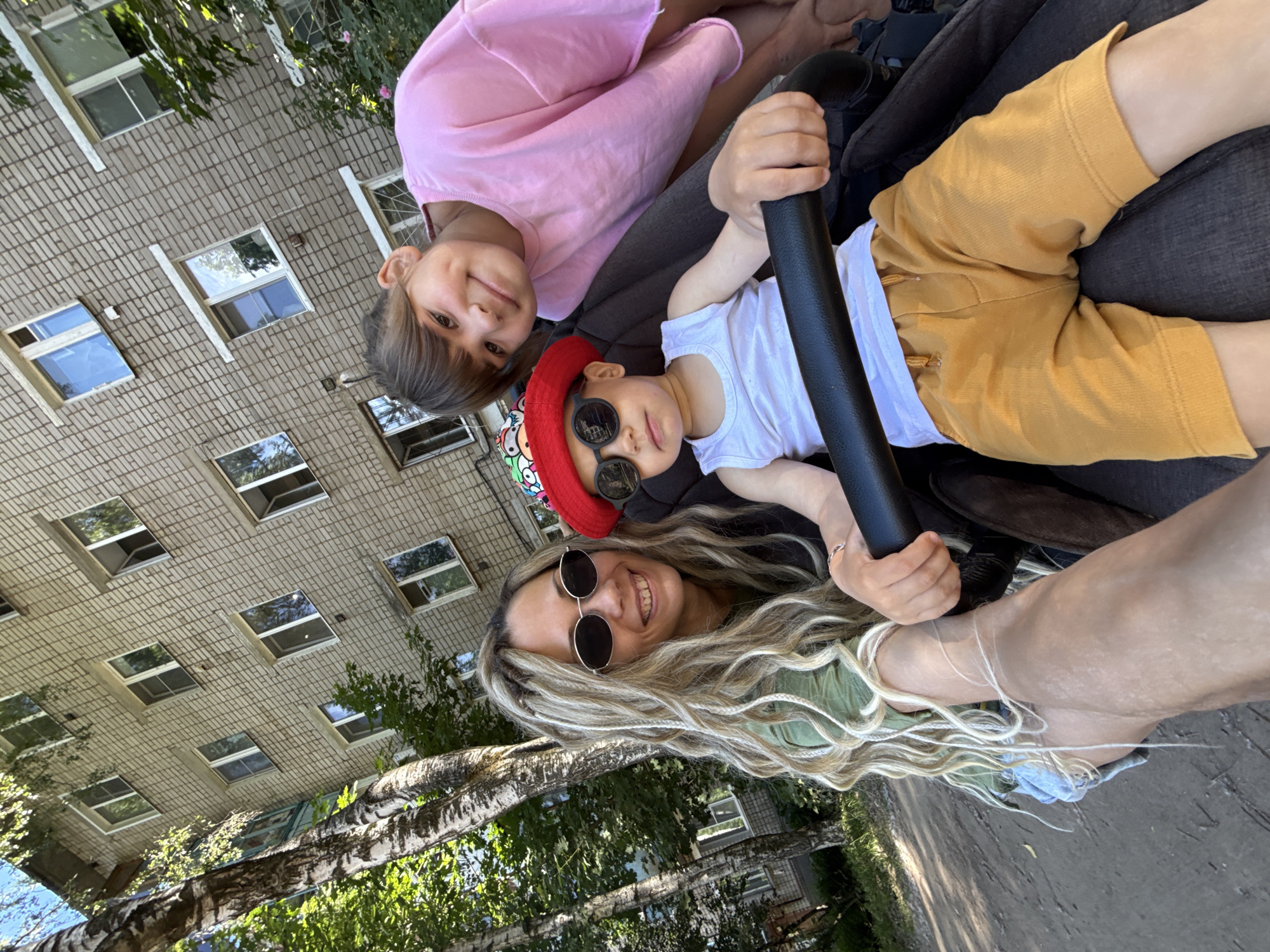 In Rusanovo, Russia, a woman with long, blonde braided hair and sunglasses is taking a selfie with two children. She is wearing a green t-shirt and denim shorts. To her right, a young boy sits in a stroller, wearing a red hat, sunglasses, and a white tank top with yellow shorts. To the woman's left and slightly behind, an older girl in a pink t-shirt smiles. They are positioned in front of a multi-story brick apartment building, with trees and sunlight casting shadows on the dirt path.