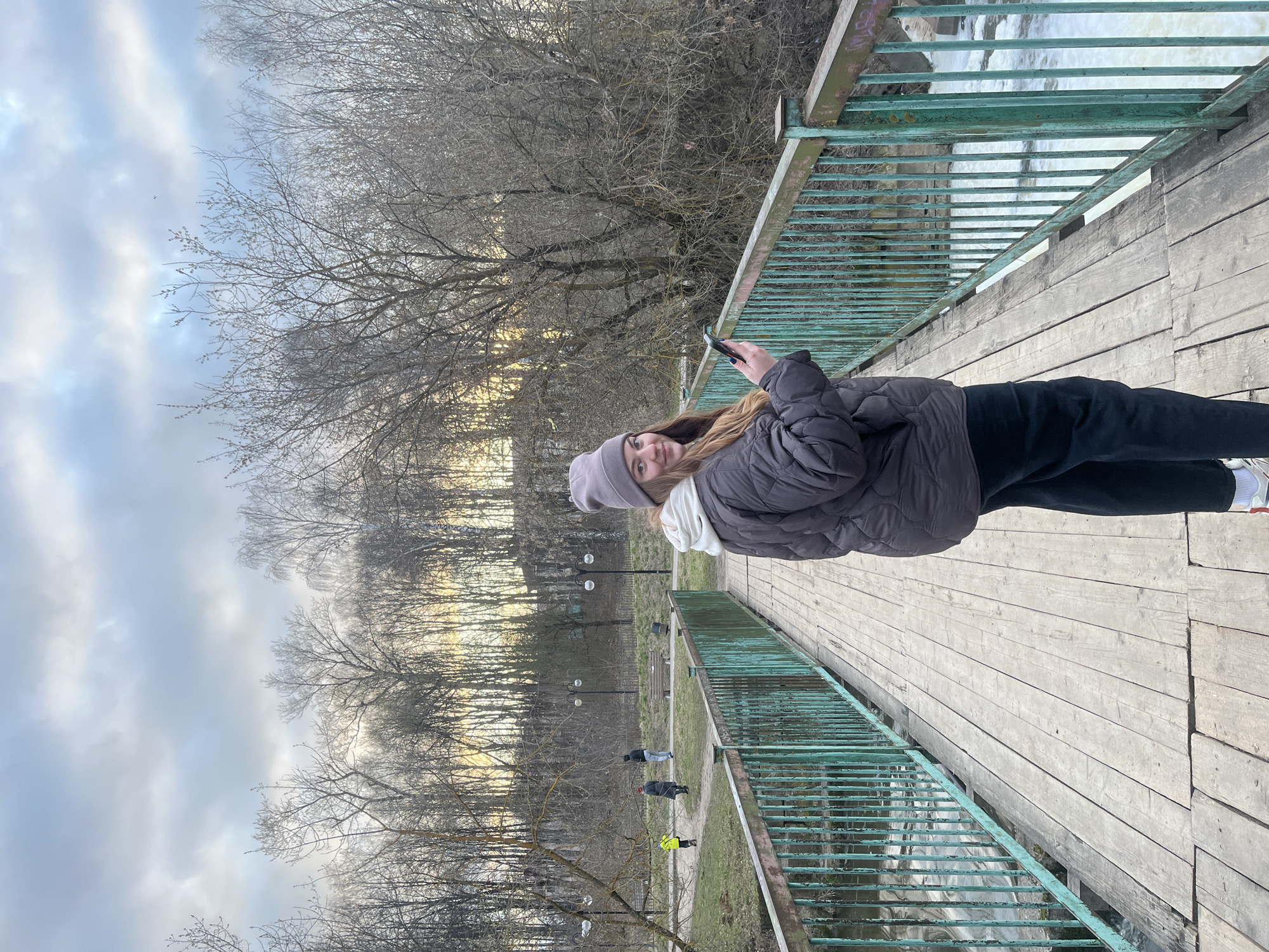 A woman wearing a grey beanie, white scarf, grey puffer jacket, and black pants stands on a wooden bridge with green metal railings, holding a mobile phone. The bridge overlooks a body of water, likely a river, with a small waterfall visible on the right side of the frame. Bare trees and cloudy skies suggest a cold or late autumn season. In the background, on the right side of the photo, are people walking in a park. The photo was taken in Klin, Russia.