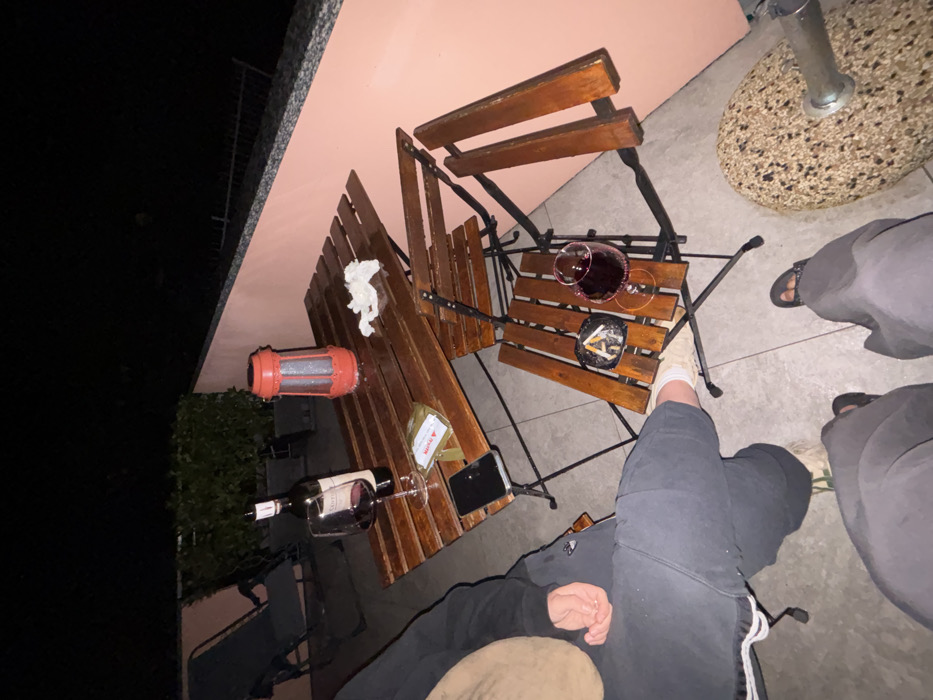 This image depicts an outdoor patio scene at night in Nesso, Italy. Two individuals are partially visible, seated or standing in the foreground. One person on the left wears dark athletic shorts or sweatpants with white trim, light-colored sneakers, and has a tan-colored object (possibly a hat or upper garment) near their head. A hand rests on their knee. The second person, at the bottom of the frame, wears dark pants and dark open-toed sandals.
The central focus is a wooden-slatted patio furniture set. A larger wooden table holds an uncorked bottle of red wine, a wine glass containing red wine, a black smartphone placed face-down, a small transparent packet labeled "MARACU," a crumpled white tissue, and a red portable lantern with a textured base. To the right, a smaller wooden-slatted chair serves as a surface, holding another wine glass with red wine and a dark circular ashtray containing multiple cigarette butts and ash. A third wooden chair, folded or partially collapsed, is positioned behind the smaller chair.
In the background, to the left, a dark grey lounge chair is visible, positioned near potted green foliage. A low, peach-colored concrete wall with a darker, speckled top edge runs along the right side of the patio. Beyond this wall, a dark, unlit area and a faint metal railing are discernible, suggesting a drop-off or lower terrace. The ground is paved with light-colored tiles. The scene is illuminated by artificial light sources, including the red lantern, against an otherwise dark night sky.
