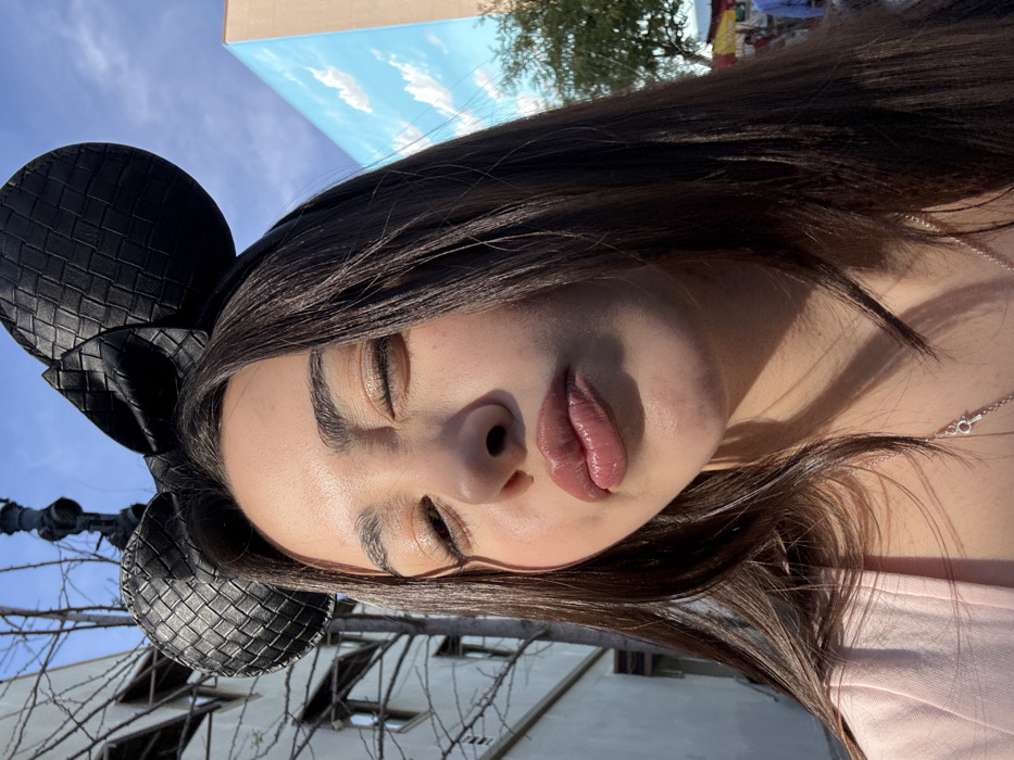 A female subject, likely a young adult, is depicted in a close-up, sunlit outdoor photograph. She has long, dark brown hair and is wearing a black, woven-textured headband featuring Mickey Mouse ears. Her right eye is closed or winking, and her lips are pursed forward, appearing glossy with a light pink tint. She wears a light pink or peach-colored garment and a silver necklace with a small key pendant.
The background to the subject's left includes a light-colored building with multiple windows and a tree with bare branches. A dark, upright pole-like structure is visible behind her head. To the subject's right, a large, bright blue wall painted with white cloud murals is prominent. Further right, green tree foliage and hints of colorful structures, possibly market stalls or umbrellas, are visible in the distance under a clear blue sky.
The setting, characterized by the Mickey Mouse ears and themed mural, is identified as the Disneyland Resort in Anaheim, California, United States of America.