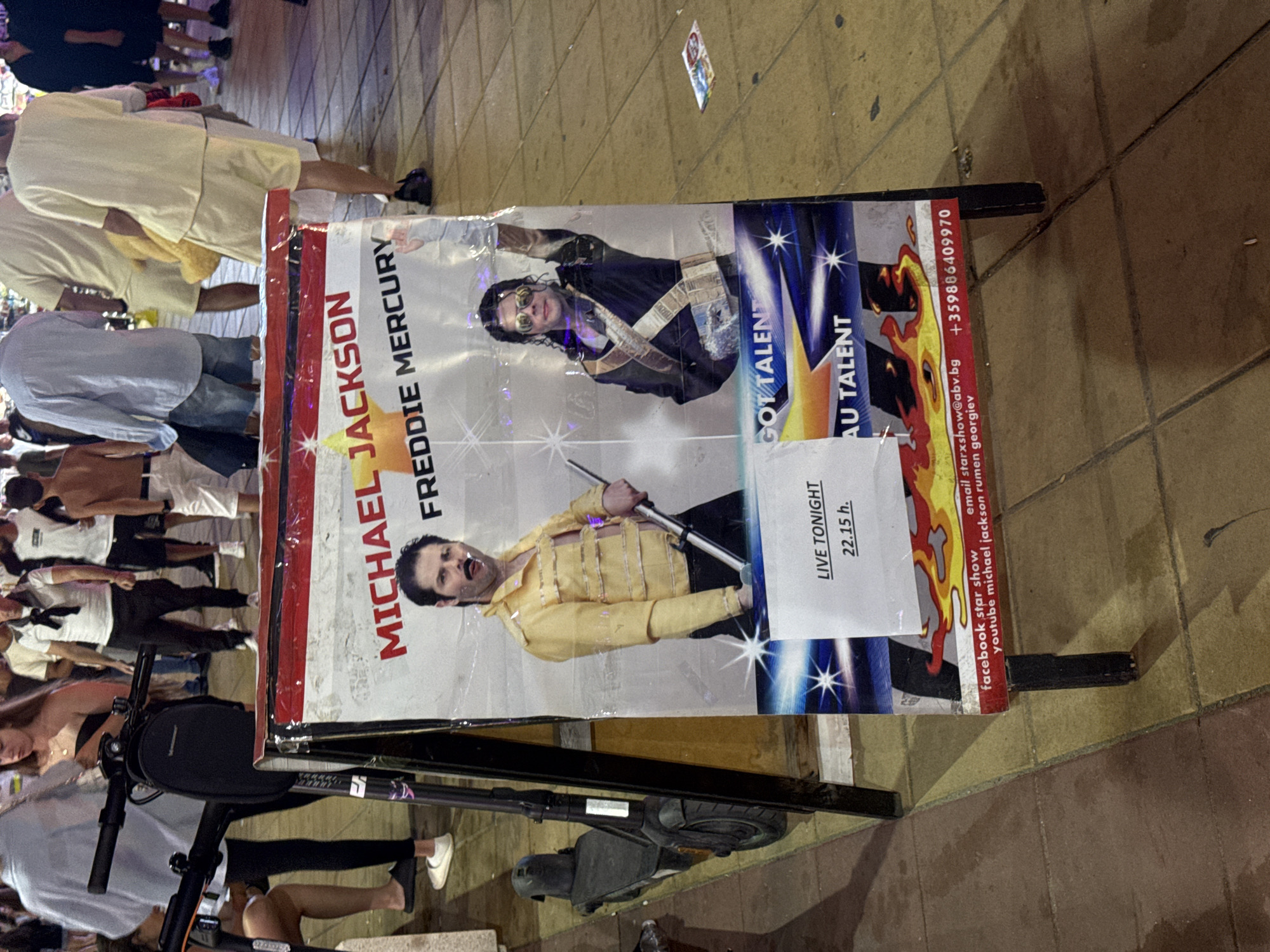 The image shows a street scene in Nesebar, Bulgaria, at night. In the foreground, there is a large A-frame sign advertising a "Michael Jackson Freddie Mercury" show. The sign features images of both performers, with "MICHAEL JACKSON" and "FREDDIE MERCURY" written in large text. Below the performer images, it states "LIVE TONIGHT 22.15 h." and mentions "GOT TALENT" and "AU TALENT". Contact information, including an email address (starxshow@abv.bg) and a phone number (+359886409970), is also visible at the bottom of the sign.
To the left of the sign, a black electric scooter is parked. In the background, several people are walking by. They are dressed in casual clothing, and some are illuminated by streetlights and the glow from the sign. The ground is paved with square tiles. The overall impression is of a busy street during an evening event.