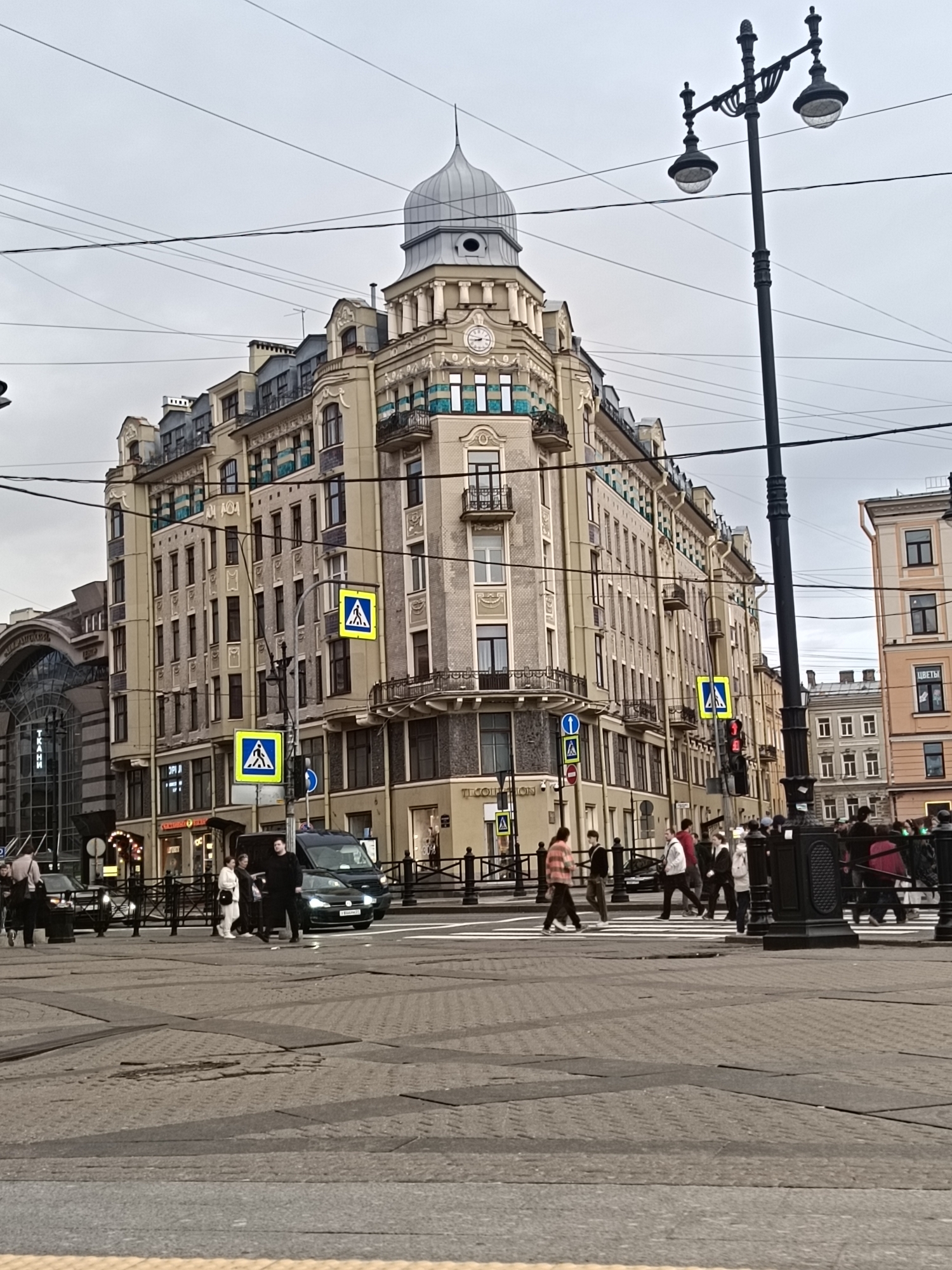 The image depicts a street scene in Saint Petersburg, Russia, showcasing a corner building with distinctive architecture, including a dome, a clock, and decorative balconies. The building appears to be located at an intersection, with pedestrians crossing the street. There are several people present, walking in different directions and waiting to cross. A dark-colored van is also visible in the traffic. Street signs and a lamp post are also present. The day is overcast. The foreground shows a paved road surface with geometric patterns.