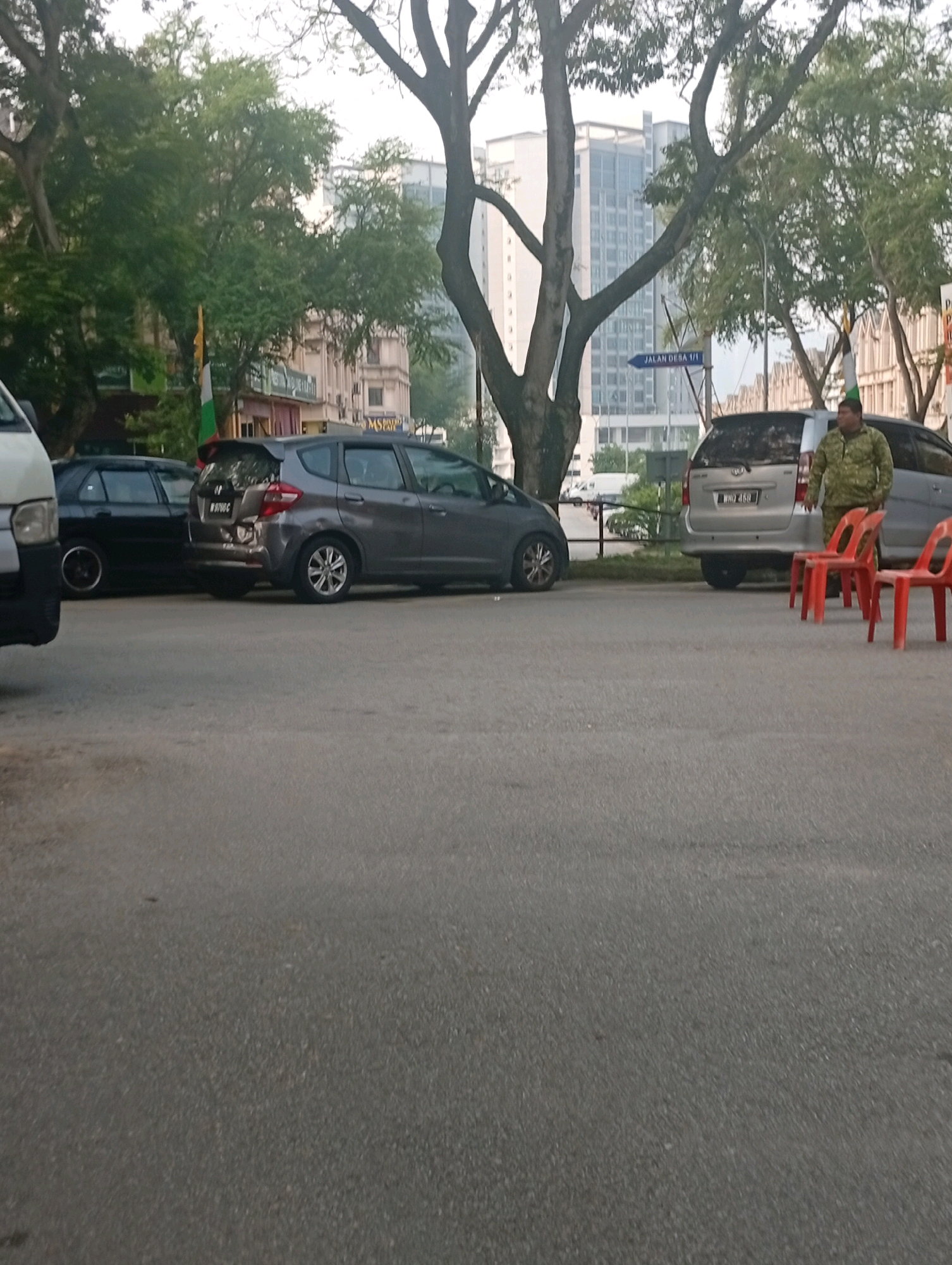 The image depicts an outdoor scene on a paved street, likely in Selayang Municipal Council, Malaysia, given the context. Several cars are parked along the roadside. In the foreground, two red plastic chairs are placed on the asphalt. To the right, a person wearing a camouflage-patterned shirt and dark pants stands near a silver van. The van has a license plate visible. Behind the cars, a variety of buildings can be seen, ranging from what appear to be shops or low-rise structures to taller, modern buildings in the background. Lush green trees with dense foliage are scattered throughout the scene, providing shade and adding to the urban greenery. A street sign with blue text indicating "JALAN DESA 1/1" is also visible, further pinpointing the location. The lighting suggests it could be daytime, possibly late afternoon or early morning, with diffused light filtering through the trees.