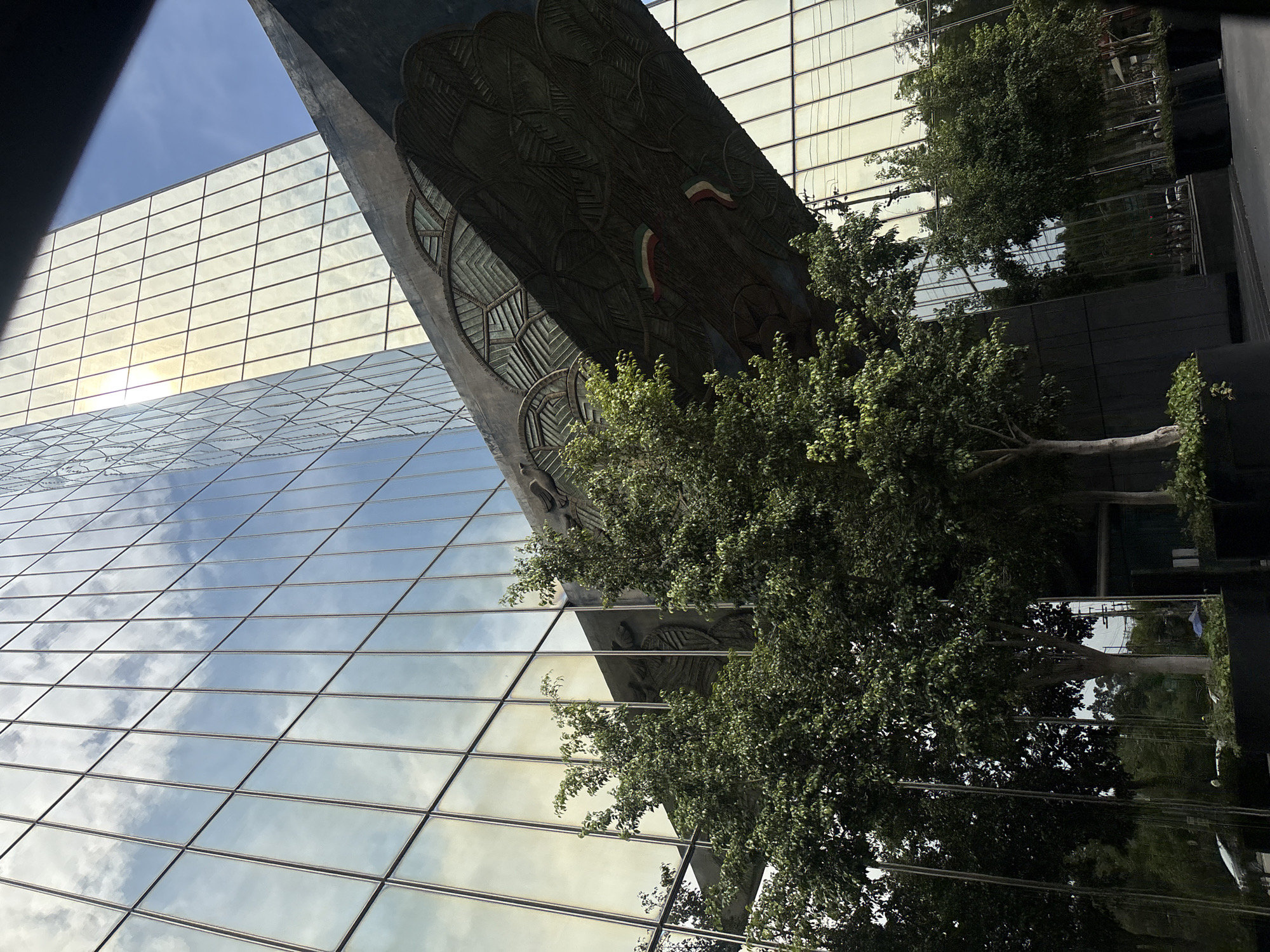 The image shows a low-angle view of a modern glass building in Mexico City, Mexico. The building's facade is composed of reflective glass panels, which mirror the sky and clouds. A section of the building features a large, sculpted stone decoration with a weathered appearance, adorned with the colors of the Mexican flag. Mature trees with dense green foliage are positioned in front of the building, partially obscuring the view. The angle and composition create an impression of towering structures and a sense of urban density.