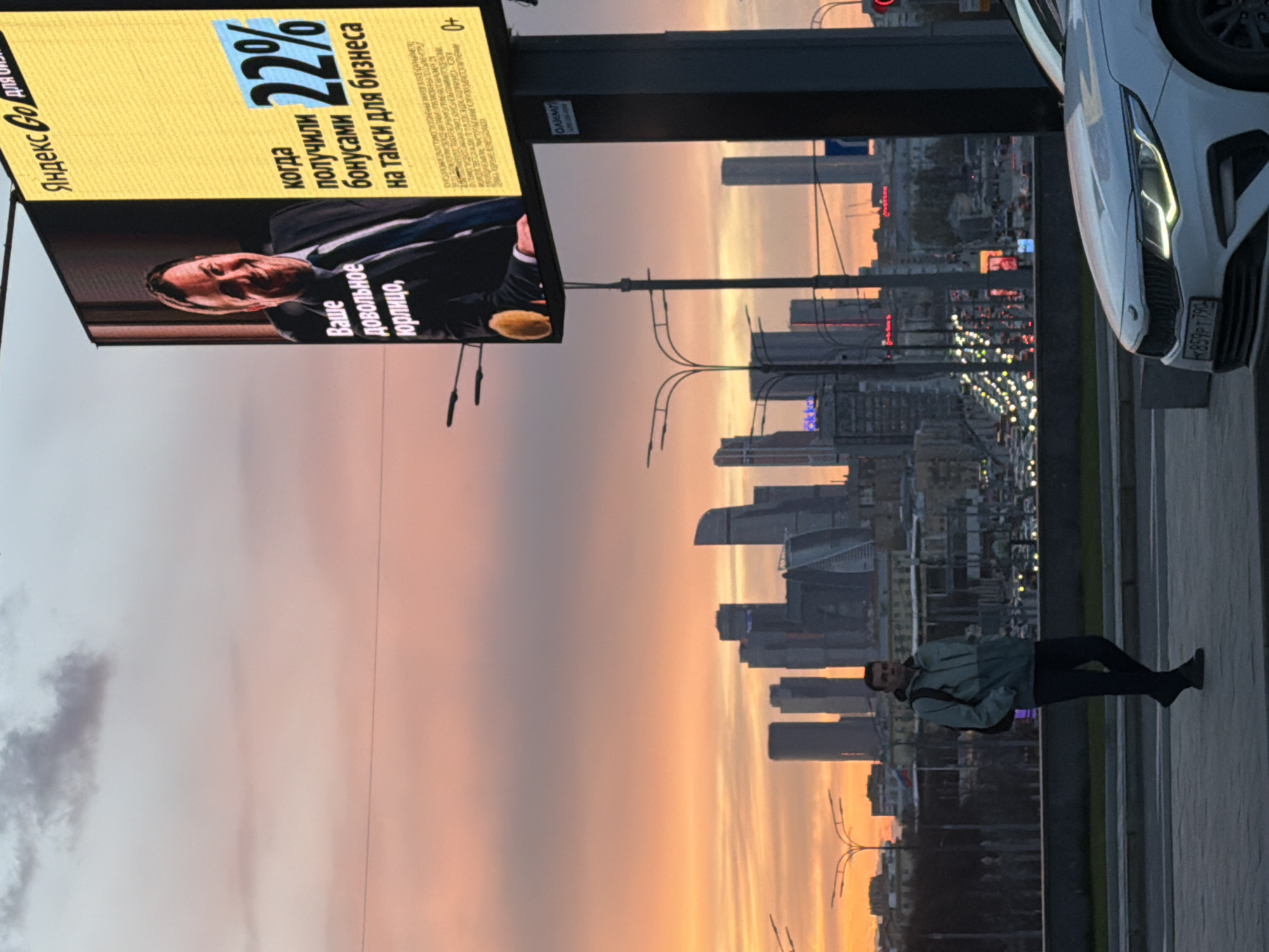 A horizontal long shot photograph taken in Moscow, Russia shows a man walking on a pathway with Moscow City's skyscrapers in the background, along with a Yandex advertisement on the left side of the photo. The sky is filled with light orange and pink hues. In the foreground on the right, part of a white car is visible. The Yandex billboard displays an advertisement featuring a man and text, including phrases in Russian.