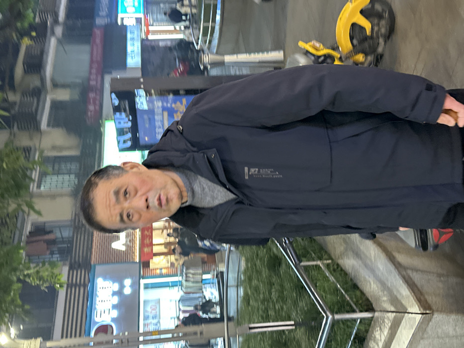 An elderly Asian male stands in the foreground of a nighttime urban street scene in Jiangjin, China. The man, centrally positioned, has dark hair, visible facial lines, and an open-mouthed expression with his eyes directed forward. He is wearing a dark navy blue hooded winter jacket over a grey turtleneck sweater; the jacket features vertical text on its left chest. To the man's immediate left, a raised planter containing green foliage is bordered by a metal railing. Behind him and to his left, a brightly illuminated storefront with blue and white signage, displaying Chinese characters and a possible English word like "EXIT," showcases clothing on racks. Further in the background, other illuminated storefronts and indistinct pedestrian figures are visible. To the man's right, a portion of a yellow shared bicycle or scooter is partially visible. The overall scene is illuminated by artificial street and storefront lighting, indicating a commercial area after dark, with multi-story buildings forming the distant background.