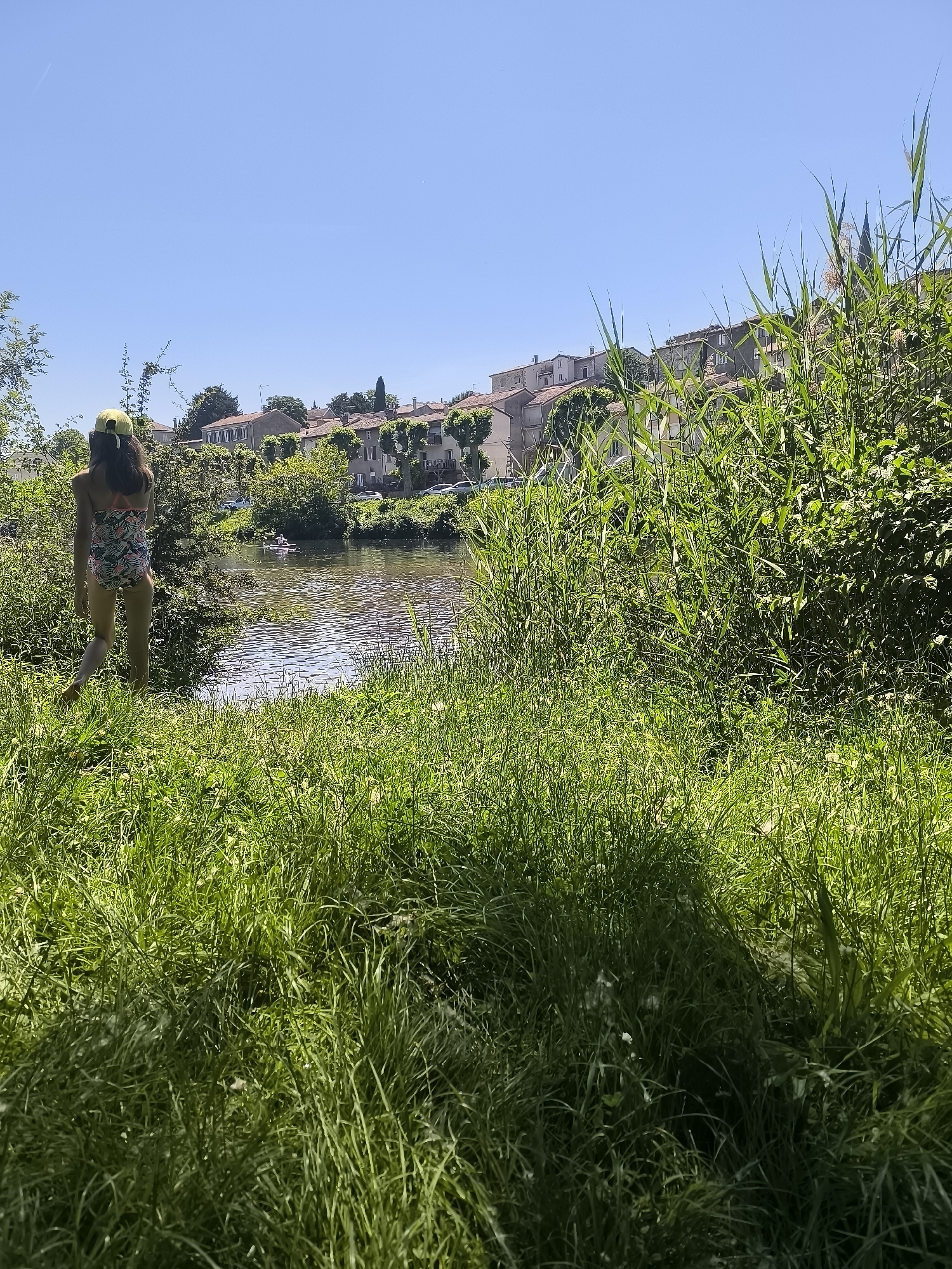 A medium shot depicts a sunny day with a young person in a swimsuit and yellow cap walking toward the Oignin river in Chézenas, France. The person is near the left edge of the frame, moving from left to right. The river reflects the clear blue sky. A row of houses with red tile roofs sits on the opposite bank. Lush green vegetation fills the foreground and right side of the frame. A small boat is visible on the river. A church spire appears behind the vegetation on the right.