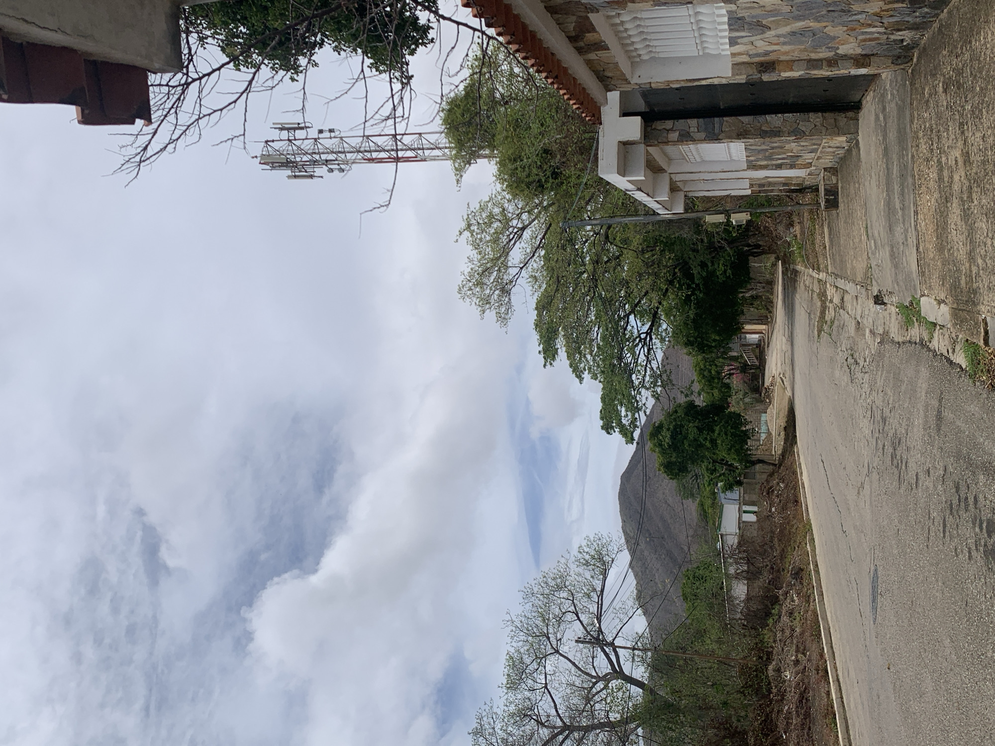 The image displays an outdoor street scene under an overcast sky, located in Santa Ana, Venezuela. The upper half of the frame shows a sky covered by extensive grey and white clouds, with occasional lighter blue patches visible between them. In the distant background, several dry, sparsely vegetated mountains are visible, with a prominent, somewhat conical peak in the center-left.
A paved asphalt road, exhibiting various cracks and a circular manhole cover in the foreground, extends from the bottom center, curving slightly left into the middle distance. On the immediate right, a concrete sidewalk with dark stains and minor debris runs alongside the road, leading to a building. This building features a facade constructed from multi-colored, irregular stone blocks (shades of brown, grey, and cream), white vertical grilles on its window openings, and a dark rectangular gate or door. Above the facade, a light-colored concrete overhang and a section of a roof with reddish-brown tiles are visible. Bare tree branches extend from the upper right, partially obscuring the sky and a red and white lattice-style cell tower equipped with multiple antennae, which is situated behind dense green foliage.
On the left side of the road, an unkempt dirt verge with dry brush and scattered refuse precedes a line of trees and bushes, some with visible bare branches. A utility pole with overhead wires is present among this vegetation. Further down the street on the left, glimpses of modest residential structures, including one with white walls and a green window frame, are discernible through the trees. No individuals or animals are visible in the scene.