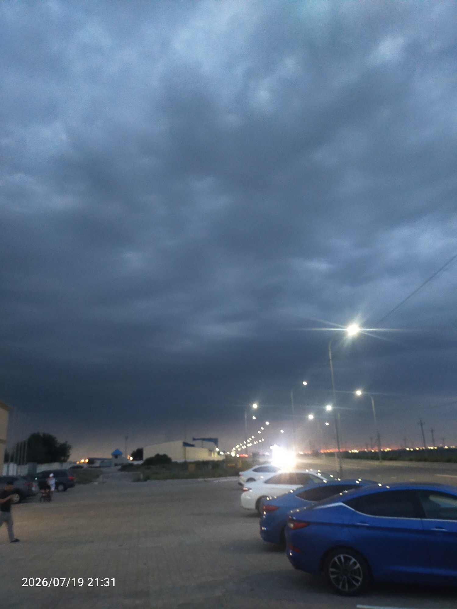 An exterior, medium shot captures a parking lot at dusk with a dramatic, dark cloud-filled sky. The sky occupies the majority of the frame, with heavy, dark gray clouds suggesting an approaching storm. Below the clouds, several streetlights are illuminated, creating bright points of light and lens flare. In the foreground, a row of cars is parked, with a bright blue sedan in the immediate foreground and two white sedans further back. The parking lot appears to be paved. In the background, buildings with illuminated windows and distant city lights are visible. The date and time stamp "2026/07/19 21:31" is present in the lower left corner of the image. There is one person visible walking in the lower left corner of the frame.