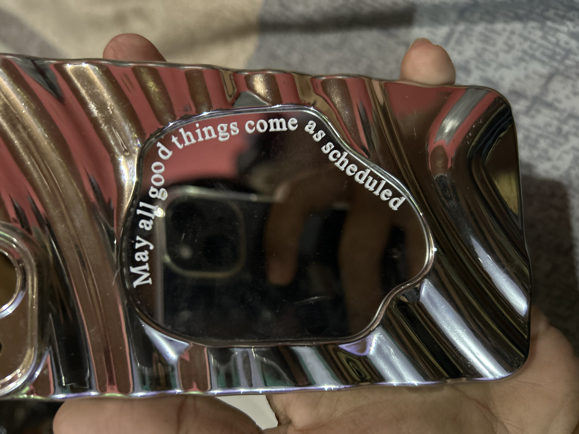 A person's hand holds a metallic phone case with a rippled, wavy texture that reflects light. In the center of the case, a rounded, mirrored surface is visible. White text is inscribed in a curve around the mirrored surface, reading: "May all good things come as scheduled". The background appears to be a textured surface, possibly fabric or carpet, in muted tones. This object is not in a recognizable location in Depok, Indonesia, as the image lacks any identifying landmarks or context.