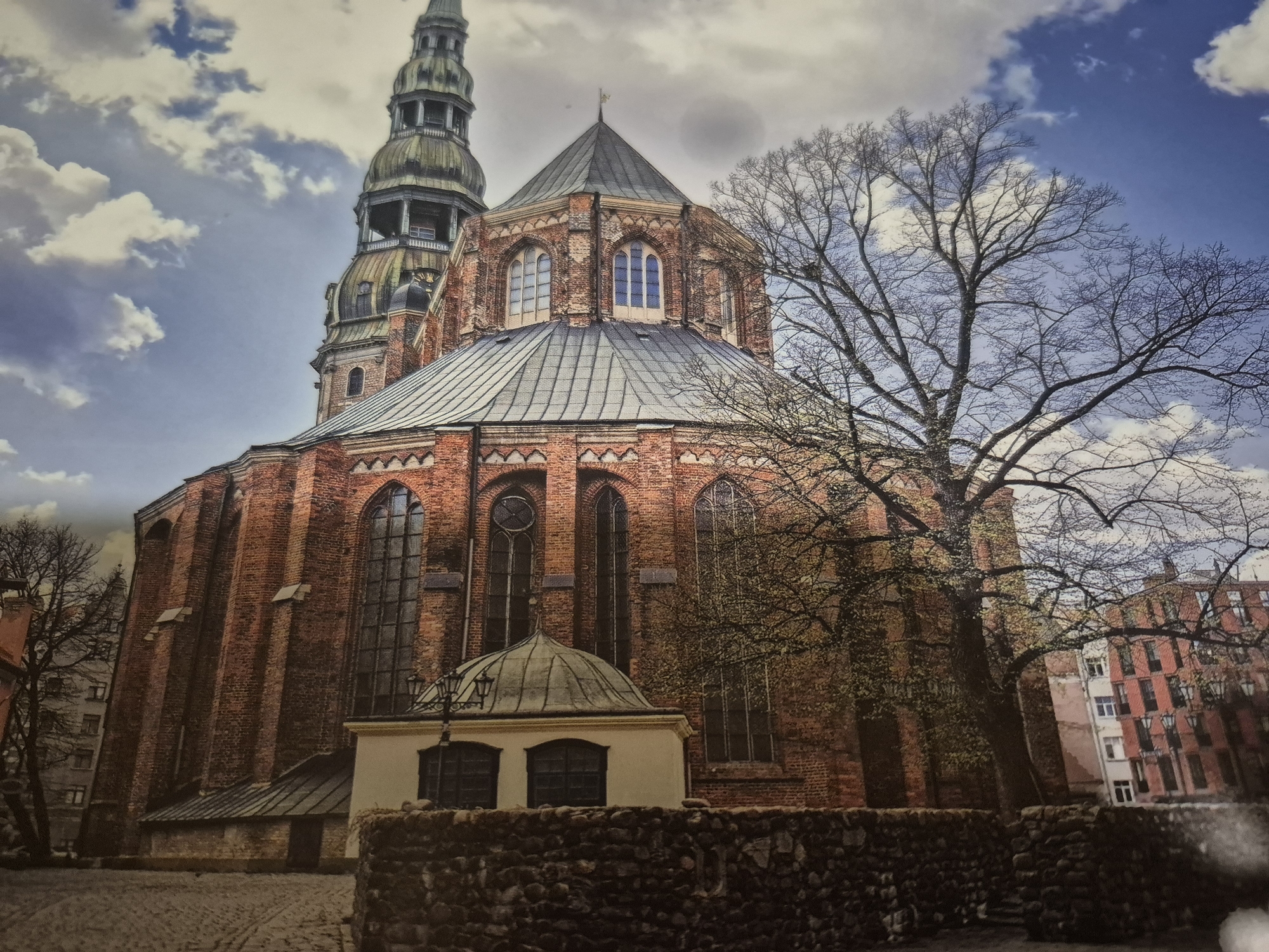 The image depicts a tall, ornate brick church with a steepled spire and a rounded, metallic roof. The church's facade features numerous arched windows, and a smaller domed structure is visible in the foreground. A bare, leafless tree stands prominently to the right of the church, its branches silhouetted against a partly cloudy sky. In the background, a multi-story building with red brick and numerous windows is partially visible. The ground in the foreground is paved with cobblestones, and a low stone wall runs horizontally across the lower portion of the image. The overall scene suggests an urban environment, possibly during a cool season given the state of the tree.