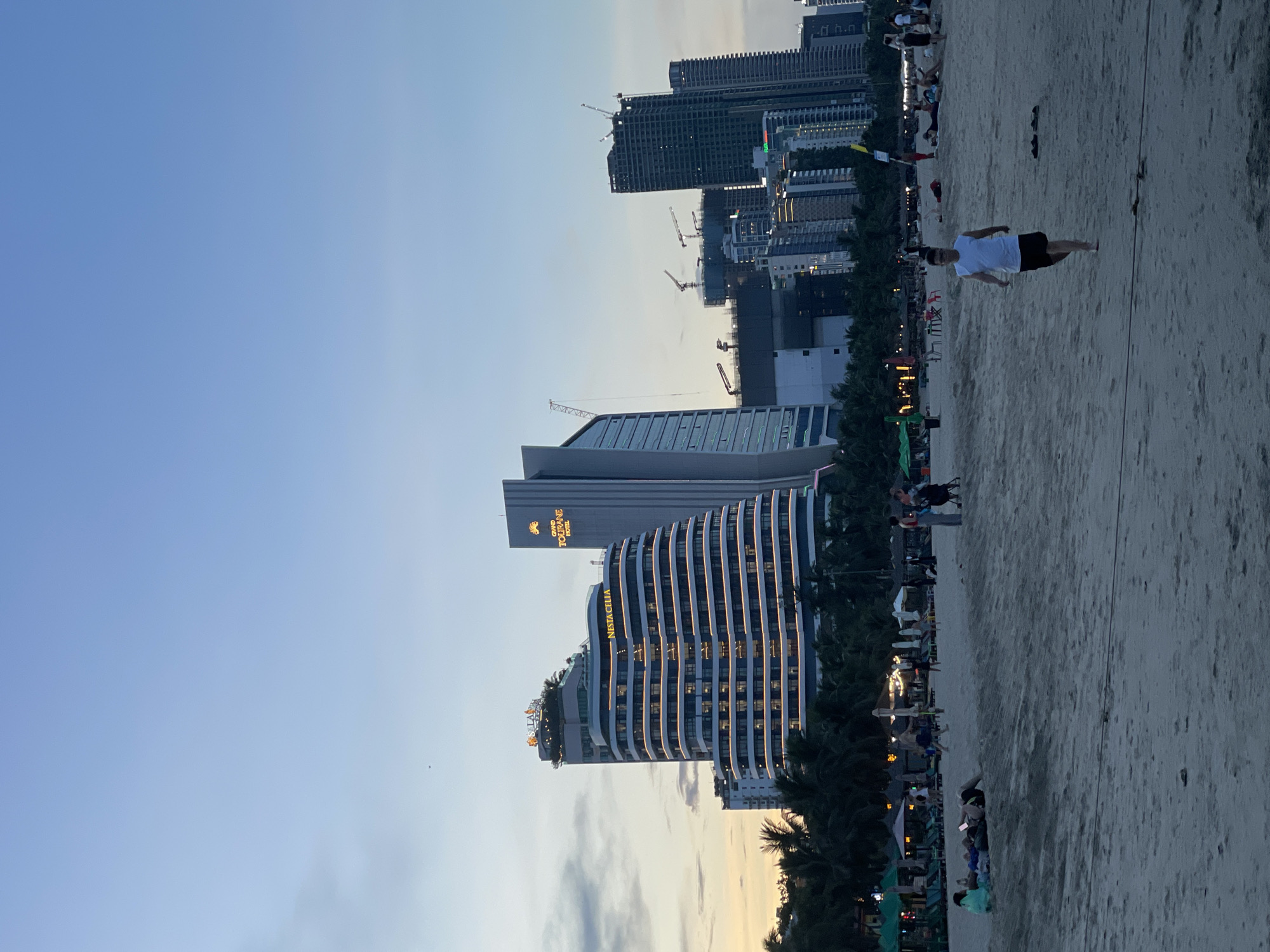 This is a wide-angle, eye-level, outdoor, full-shot photograph taken on a beach in Da Nang, Vietnam, at dusk. The sky is a clear gradient of light blue to pale grey, with a few wispy clouds near the horizon. In the background, a modern cityscape rises behind a line of palm trees. Prominent buildings include the curved, illuminated hotel NESTA CELIA and the GRAND TOURANE HOTEL. Several other high-rise buildings, some under construction with visible cranes, dominate the skyline.
The beach in the foreground is sandy, with scattered footprints and some debris. In the middle ground, a man in a white sleeveless shirt and black shorts walks towards the camera, barefoot. Further back, several beach umbrellas, lounge chairs, and a few people are visible, some relaxing on the sand or walking along the shore. The overall scene depicts a relaxed evening atmosphere on a popular beach in Da Nang.