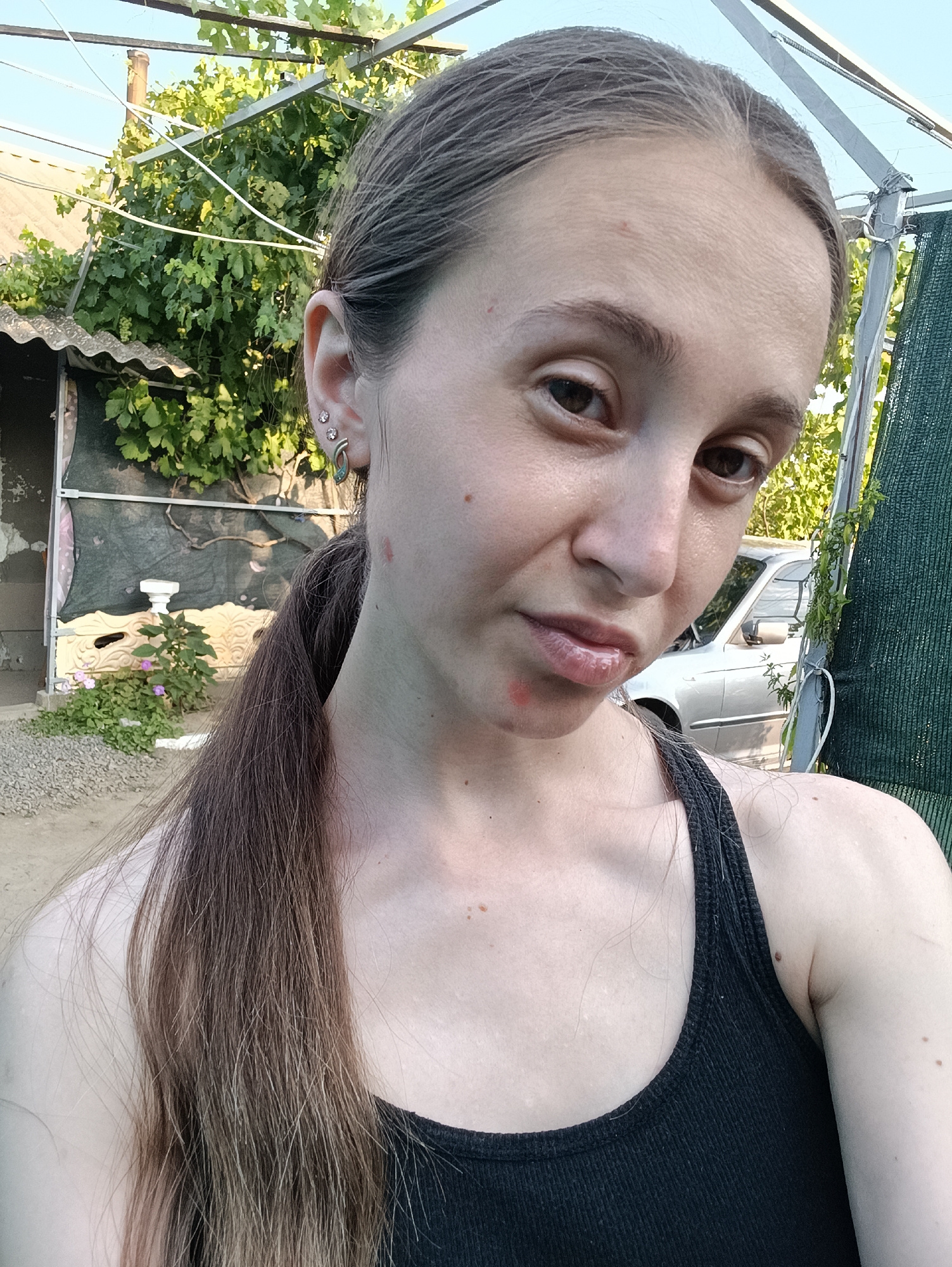 A young woman with light brown hair pulled into a ponytail is taking a selfie outdoors. She is wearing a black tank top and has several piercings in her left ear. Her skin appears to have some redness on her face and neck. She is standing in what appears to be a backyard or outdoor area, with a grape vine overhead, a corrugated metal shed, and a car visible in the background. The location is identified as Vypasne, Ukraine. The overall lighting suggests it is daytime.