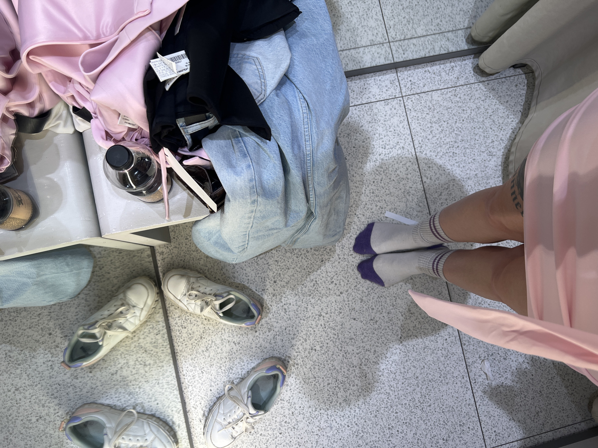 The scene depicts the interior of a retail fitting room in Riga, Latvia, viewed from a high-angle perspective. The floor is a light gray terrazzo or speckled tile surface with visible grout lines. A large mirror occupies the left side of the frame, reflecting the floor and adjacent items.
The lower legs and feet of one individual are visible at the bottom center. The person is wearing white crew socks with purple cuffs and thin purple stripes near the ankles. A light pink garment drapes over the individual's knees. A tattoo is partially visible on the person's left upper thigh/hip area.
On the tiled floor to the left, a pair of white athletic shoes with light blue and pink accents is placed. These shoes are also visible as reflections in the mirror.
To the right, a pile of clothing and objects rests on a light-colored shelf or bench. This pile consists of light-wash blue denim jeans, a quantity of light pink fabric, and dark fabric with a visible white retail tag. A clear plastic bottle with a black screw-top cap, containing a dark liquid, is positioned on the shelf amongst the clothes. Partially visible are black-framed sunglasses and a white tag. A light-colored fabric curtain or panel is visible along the far right edge of the frame, indicating the boundary of the fitting room enclosure.