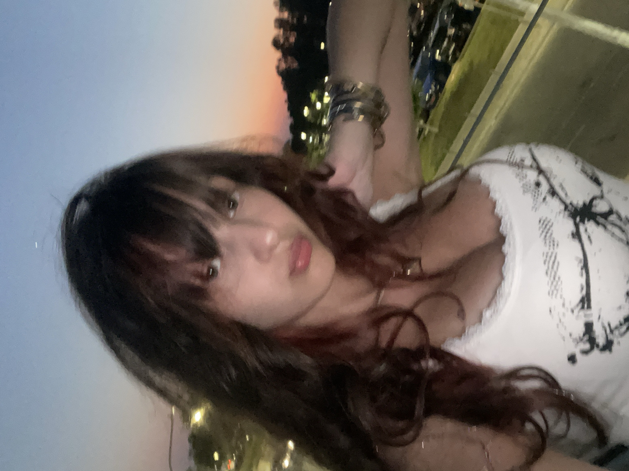 A young woman with long, dark, wavy hair and bangs is pictured from the chest up. She is wearing a white tank top with a black graphic print and a lace trim. She has several bracelets on her left wrist. The background shows a dusky sky with hints of orange and pink, suggesting sunset or sunrise. Beyond that, there are silhouetted trees and a few streetlights, indicating an outdoor, possibly urban or suburban setting at dusk. The image is taken from a slightly elevated perspective, looking down towards the scene. The location is Paphos Municipality, Cyprus, possibly near a coastal road or a viewpoint overlooking a parking area.