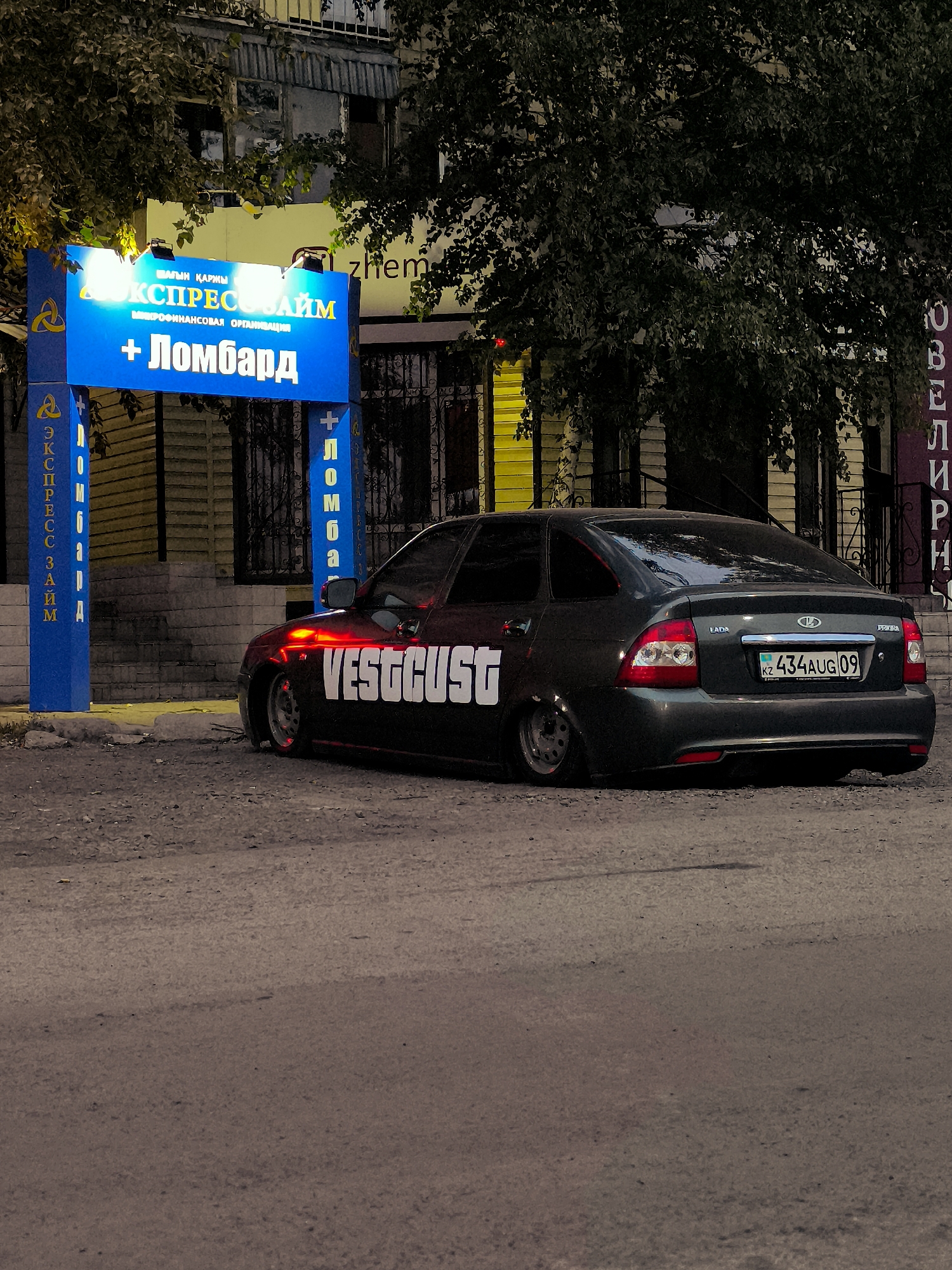 A dark-colored sedan is parked on a gravelly surface in front of a building with a bright blue sign. The sign, illuminated at night, features text in what appears to be Cyrillic and Latin alphabets. The building behind the sign has windows and is partially obscured by trees. The car has a custom decal on its side that reads "VESTGUST" in large white letters. The license plate of the car is visible and reads "KZ 434AUG09". The overall scene suggests an urban environment at night.