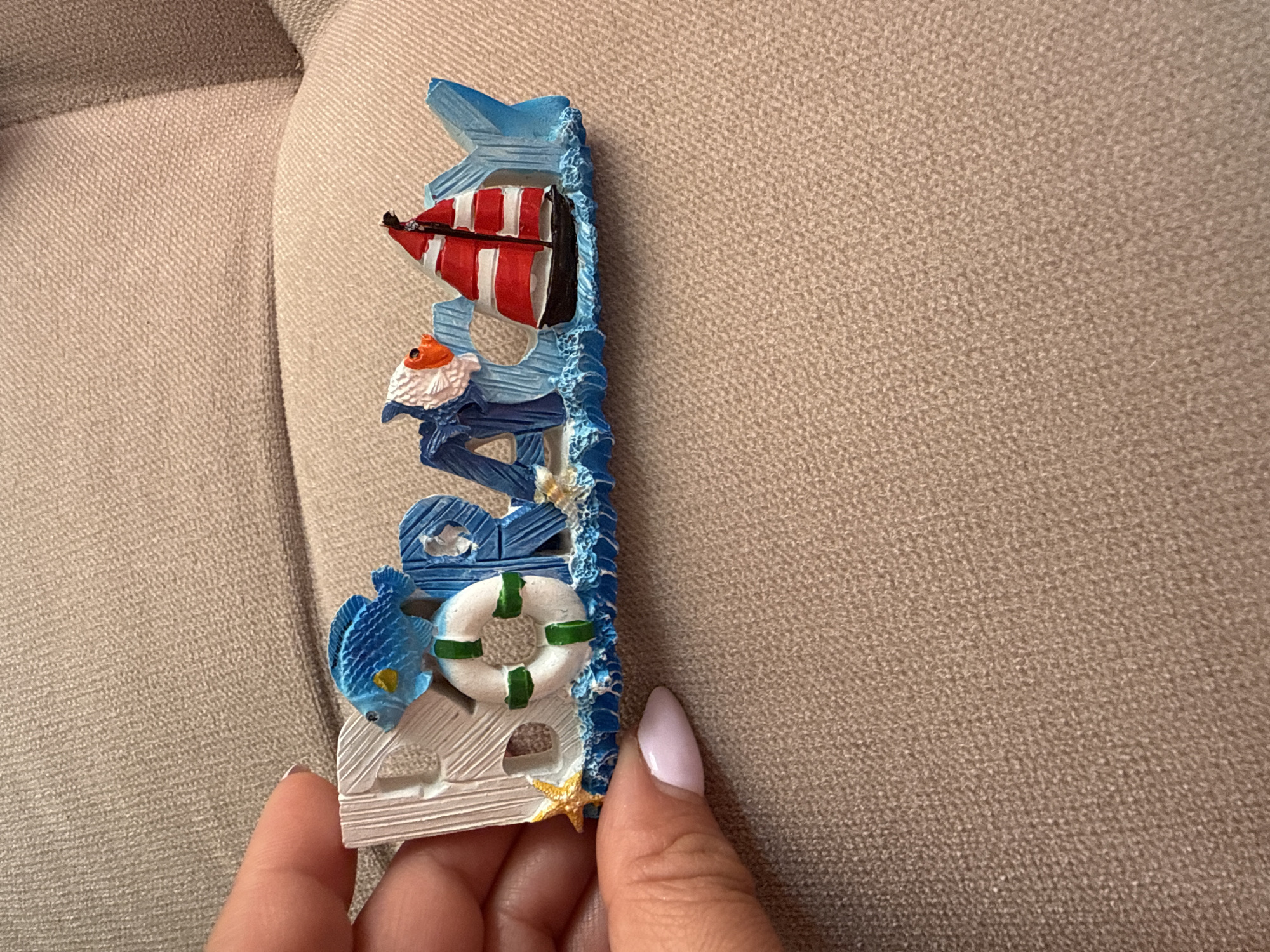 A close-up, eye-level shot captures a person's hand holding a vertical, nautical-themed decorative object against a textured light beige fabric background, likely a sofa or chair.
The colorful decorative object, possibly a souvenir or magnet, features various marine elements stacked vertically. At the top, a small sailboat with distinct red and white striped sails is depicted. Below the sailboat, a small figure resembling a fisherman or gnome with an orange hat and a white beard is visible. Further down, partial blue letters spelling "SEA" are integrated into the design. To the left of these letters, a blue fish is shown, and beneath it, a white lifebuoy with green stripes. The base of the object is designed to look like white wooden planks, accented by a small yellow starfish. The entire right edge of the object is bordered by textured blue elements, resembling ocean waves.
A hand, featuring light pink manicured nails on the thumb and index finger, holds the bottom part of the decorative item. The background fabric displays a finely woven, slightly bumpy texture, appearing uniform in its light beige-brown hue across the frame. The scene is brightly and evenly lit, suggesting an indoor environment. No other activity is observed, and there are no explicit cues for location, time of day, or specific emotions.