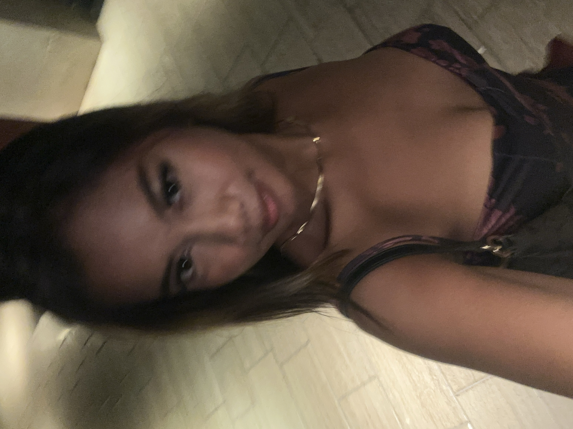 This slightly blurry and grainy close-up image features a young woman posing for what appears to be a selfie, with her face tilted towards the camera from the upper left side of the frame. She has long, dark hair and gazes directly at the viewer with a gentle, subtle smile. A delicate gold-colored chain necklace is visible around her neck. She is wearing a dark-colored top or dress, which features a faint floral or abstract pattern in shades of purple or deep red with lighter accents.
The background is dimly lit and out of focus, showing light-colored, possibly tiled or plank flooring on the left and bottom, and indistinct, light-toned surfaces, perhaps walls or furniture, in other areas. The dim lighting conditions suggest the photo was taken indoors during the evening or night. The angled composition and close proximity indicate a self-portrait. There is no visible text in the image.