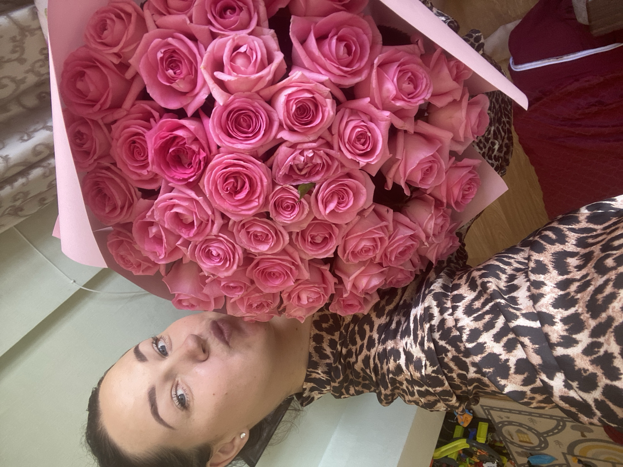 The image shows a woman holding a large bouquet of pink roses. She is wearing a leopard-print top. The background includes parts of a room with items like curtains, bedding, and a child's play area. The location, city of Sochi, Russia, is not discernible from the visual information in the image.