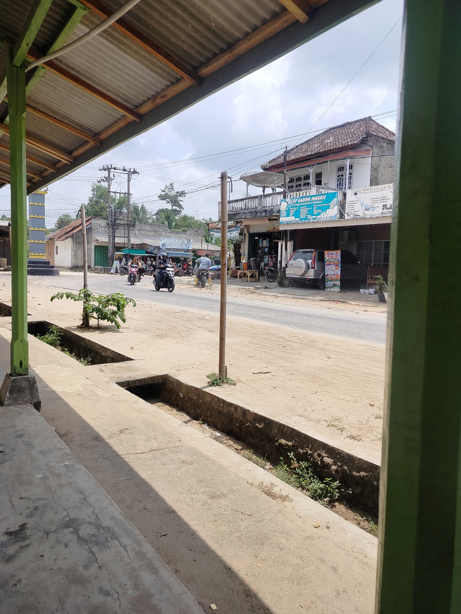 This is a medium-shot, eye-level, outdoor, slightly overcast image captured from a covered porch looking out onto a dusty road in Sri Gading, Indonesia. The foreground is dominated by a concrete floor with a drainage ditch running alongside it. The ditch contains some sparse vegetation and dirt.
Beyond the ditch, a paved road stretches into the distance, with several motorbikes visible. One motorbike, ridden by a person wearing a helmet, is in the left lane. To its right, another motorbike, also with a rider, is in the right lane. Further down the road, a third motorbike with a rider wearing yellow pants is visible. A silver SUV is parked on the right side of the road in front of a building.
The buildings lining the road appear to be a mix of shops and residential structures. One building has a sign in English reading "LRP GADING INGGRIS". Another sign, also in English, is visible on a stand in front of the silver SUV. Electrical poles and wires crisscross the sky. A tall, patterned pillar with yellow and blue bands is visible on the far left of the image. The overall scene depicts a typical street in a local Indonesian town.
