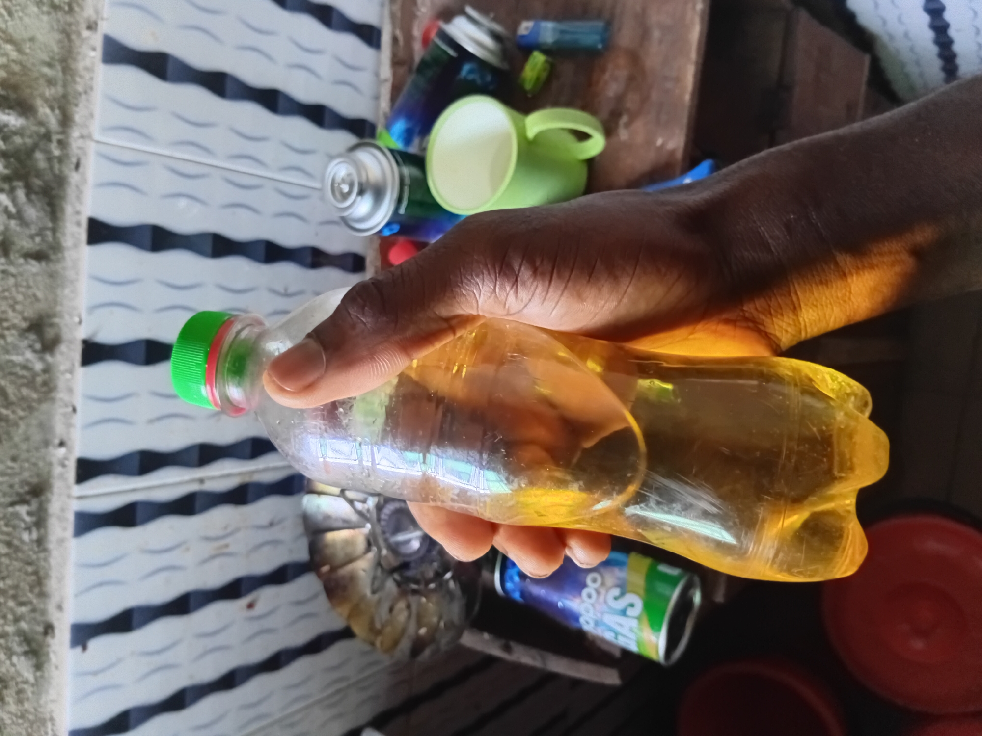 The image shows a person holding a transparent plastic bottle filled with a yellow liquid. The bottle has a green and red cap. The person's hand is dark-skinned. In the background, there are several objects including aerosol cans, a green plastic cup, a blue lighter, a metal object, and a can with green and white label. A red plastic bowl is also visible in the background. The background includes a tiled wall. The location is identified as Akwete, Nigeria.