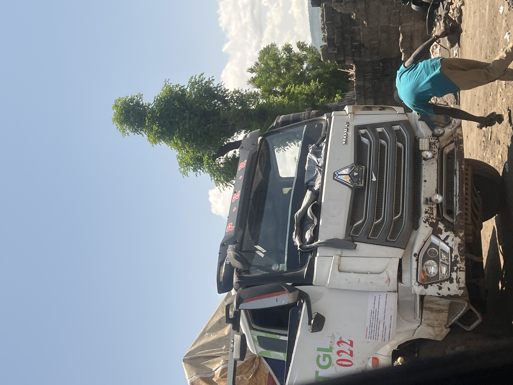 The scene depicts a white Sinotruk HOWO heavy-duty truck parked outdoors in Kwarge, Nigeria. The truck's front bumper is visibly damaged with extensive paint peeling and scuff marks. Headlights are present and appear functional. On the passenger side of the truck's cab, a green "TGL" logo with the red number "022" is affixed. Below this, a white paper sign is visible, identifying the vehicle as operating "exclusively on consignment with KOTTO FREIDA LIMITED, located in Saminaka LGA, Fadan Kaje." The roof-mounted sun visor bears partially visible red lettering, "PRINCE," and Chinese characters. A light brown tarp covers cargo in the rear section of the truck, which is partially visible.
A single adult male is present, positioned at the front-right side of the truck (from the viewer's perspective, or the driver's side from the truck's perspective). He is wearing a bright turquoise short-sleeved t-shirt and light brown pants, and is bent over, facing away from the camera. His arms are extended towards the truck's bumper area, holding a dark object in his left hand, suggesting an act of inspection, cleaning, or minor repair.
The ground surface is unpaved, consisting of dry, reddish-brown dirt. The background features a low wall constructed from rough-hewn stones or concrete blocks, beyond which several large green trees with dense foliage are visible. The sky is light blue with scattered white cumulus clouds, indicating clear weather conditions. Distant hills or elevated terrain are faintly visible on the horizon.
