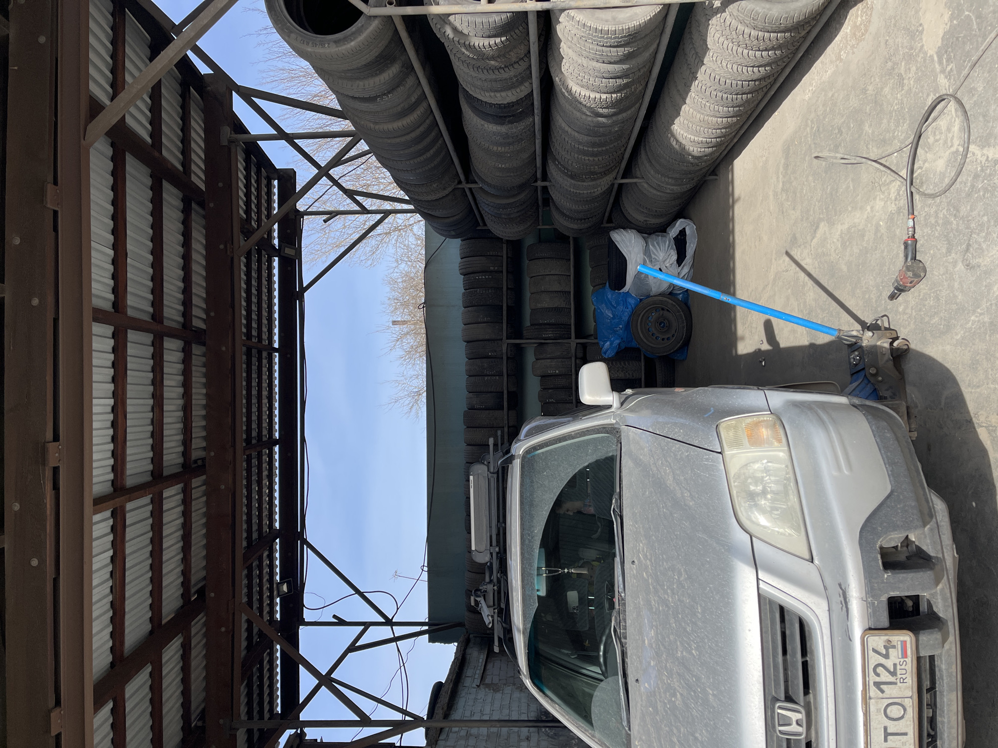 The image depicts a silver Honda CR-V parked in what appears to be a tire shop or garage. Stacks of tires are arranged on shelves along the wall. The vehicle has a license plate, which appears to read "TO 124 RUS" and indicates the location is in Russia. To the right of the vehicle are tools including a jack with a blue handle and an air powered impact wrench. Overhead is a metallic structure with a corrugated roof. Based on the license plate, the location is in Russia, specifically the 124 region, which corresponds to the city of Krasnoyarsk.