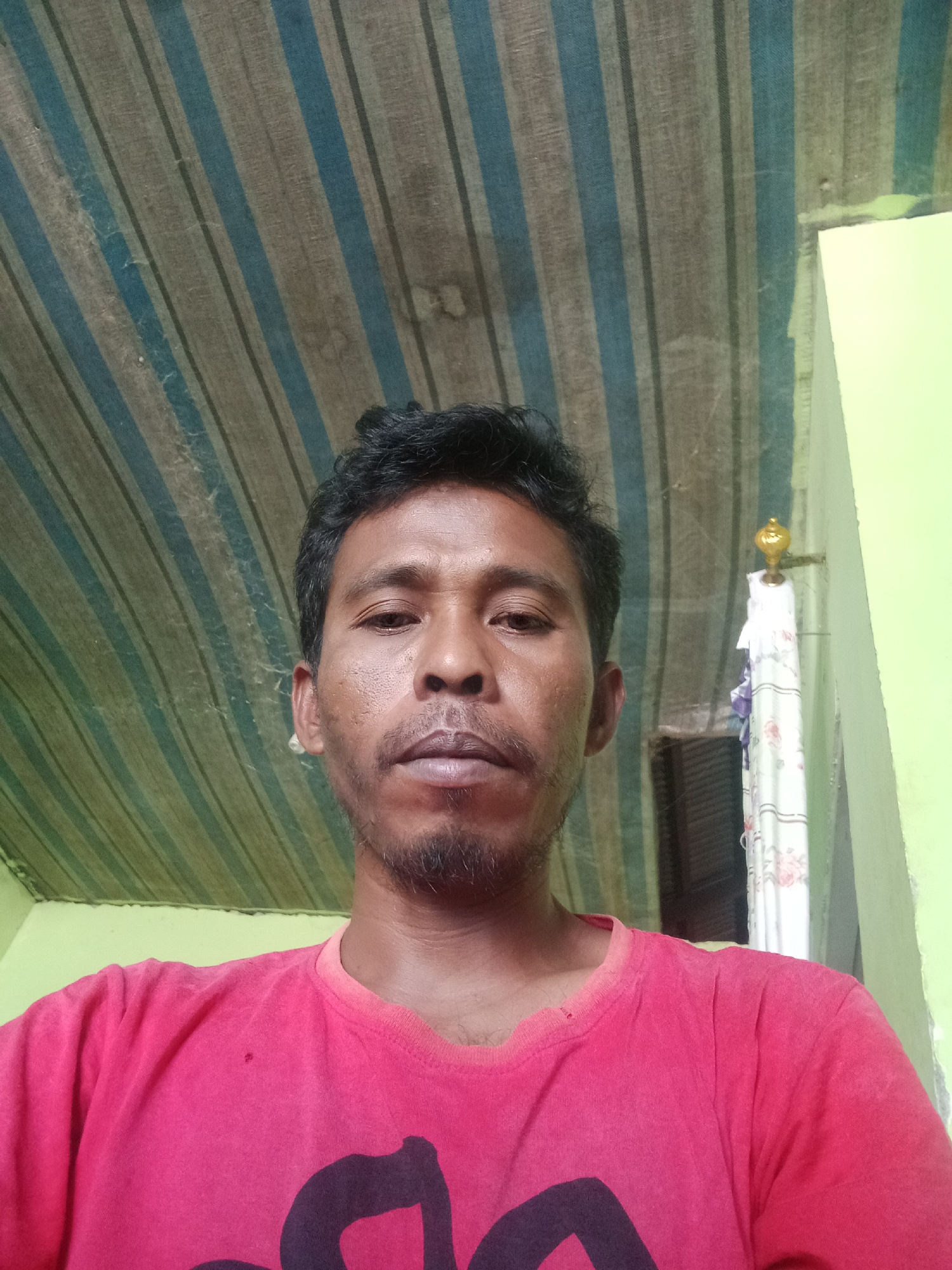 A man, appearing to be of Southeast Asian descent, is photographed from the chest up, looking directly at the camera. He has dark, curly hair and a goatee. He is wearing a bright pink t-shirt. The background consists of a striped tarp overhead and a light green wall to the right. A decorative hook with fabric hanging from it is visible on the wall. The overall impression is that of a candid self-portrait taken indoors or in a covered outdoor space. The location is identified as Lakea Satu, Indonesia.