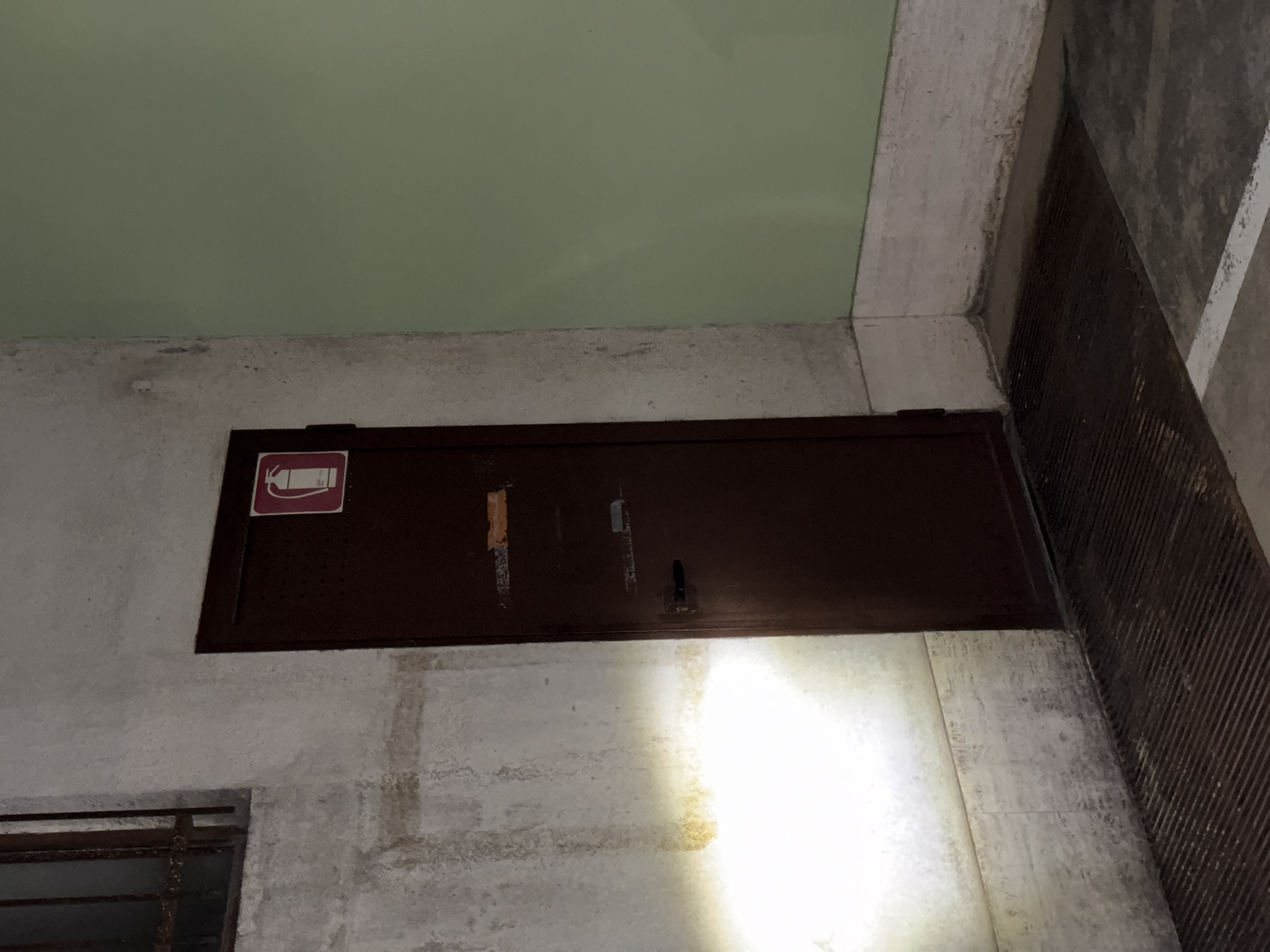 The image shows a low-angle view of a dark brown, rectangular access panel set into a concrete wall. The panel has a red and white fire extinguisher symbol on its left side. Above the wall is a green ceiling with a concrete border. To the right of the wall, there's a section of fluted, possibly metal or concrete, material. The lighting in the lower-right of the image is bright, possibly from a flashlight. The lower-left corner shows part of a metal-barred window or vent. Location: Unknown, Unknown.