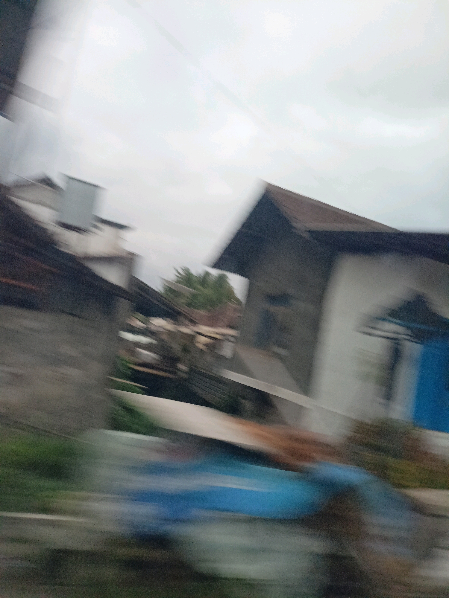 The image depicts a blurred scene of buildings and vegetation, suggesting motion or a low-quality camera. In the foreground, a blue object, possibly a tarp or fabric, is visible. Behind it, a grey wall with some texture and greenery are discernible. Further back, a house with a dark, angled roof and white walls is present. To the left, another building with a dark roof and grey walls is seen. The sky is overcast and grey. The scene appears to be a residential area, possibly in Payaman, Indonesia, given the architectural style and vegetation. No people or specific actions are clearly identifiable due to the blurriness.
