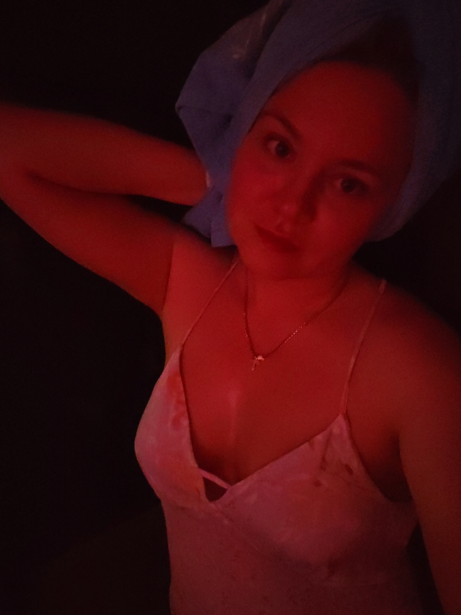 In a dimly lit room, a young woman with a blue towel wrapped around her head and wearing a pink negligee is captured in a selfie. The red lighting casts a strong glow across her face and upper body. She is looking directly at the camera, and her right arm is raised, touching the towel on her head. A delicate necklace with a small pendant adorns her neck. The background is dark, with only a hint of her shoulder and arm visible on the left side of the frame. The scene appears to be a private indoor setting, possibly a bathroom or bedroom, within an apartment in Izhevsk, Russia.