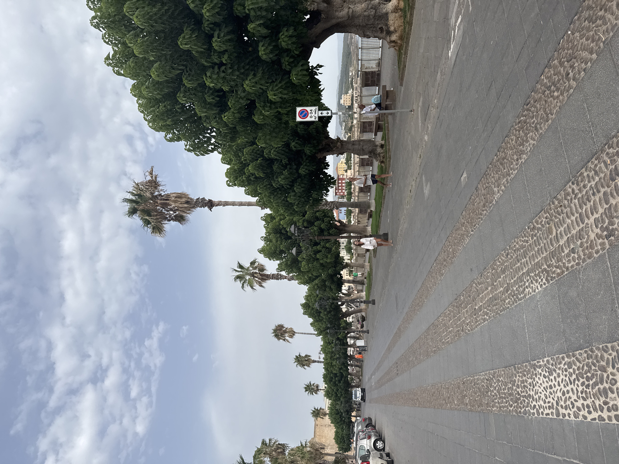 The image depicts a wide, paved promenade along a waterfront in Alghero, Italy. The promenade is lined with a row of mature trees, some of which are palm trees, casting shadows on the pedestrian walkway. The walkway itself is paved with large rectangular stones, interspersed with decorative bands of small, rounded pebbles.
On the left side of the promenade, a line of parked cars is visible, suggesting a public parking area. In the distance, buildings and other elements of the cityscape can be seen, including what appears to be a historic stone wall.
Several people are present on the promenade. A woman in white shorts and a white top is walking away from the camera on the right side. Further along, a man in a white t-shirt and blue shorts is walking towards the camera. Another man is seated on a bench on the right side, with a blue bag next to him. A few other individuals are visible in the background, engaged in casual activity.
A traffic sign indicating parking restrictions (0-24) is posted on the right side of the promenade. The sky is partly cloudy, with a mix of white clouds and blue sky. The overall impression is a pleasant, public space by the sea, likely a popular area for strolling and recreation in Alghero.
