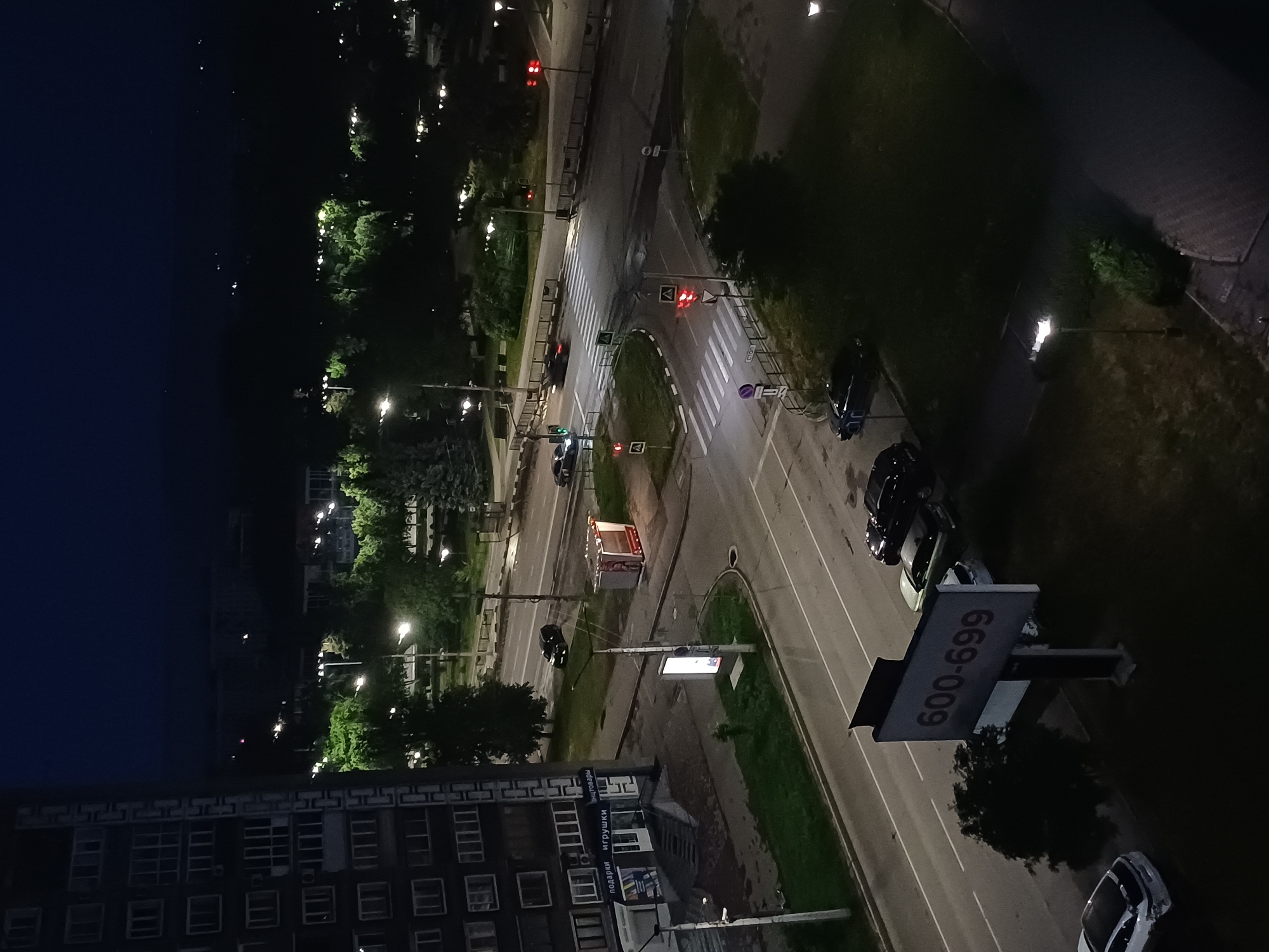 The image is a nighttime aerial view of a city street intersection. On the left is a large, multi-story apartment building with many windows. Across the street, there are trees and streetlights illuminating the area. The intersection itself has pedestrian crosswalks and traffic lights. Several cars are visible on the road, some parked and some in motion. A bus stop shelter with a red frame is present near the intersection. In the foreground, to the right of the apartment building, is a large billboard displaying the numbers "600-699". The sky is dark, indicating it is nighttime, and there is some ambient light from streetlights and building windows.